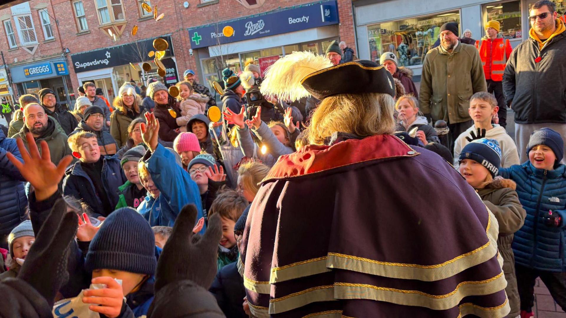 Children in the street in Driffield with their arms raised as sweets are thrown in the air. The spectacle is being watched by a group of adults and local dignitaries