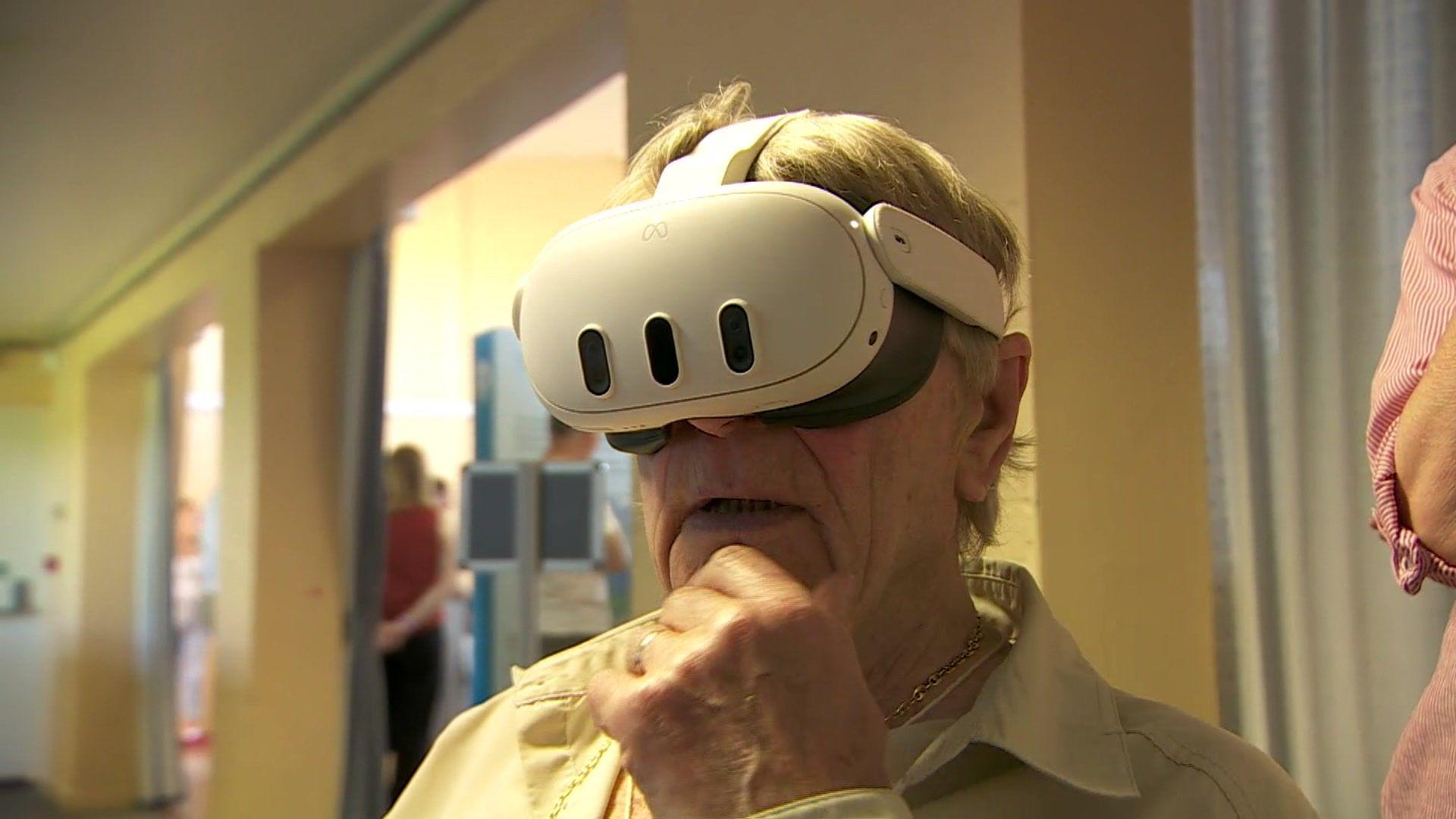 A man with grey hair is wearing a white virtual reality set. His hand is on his  chin, reflecting he ponders what he sees and his mouth is slightly ajar