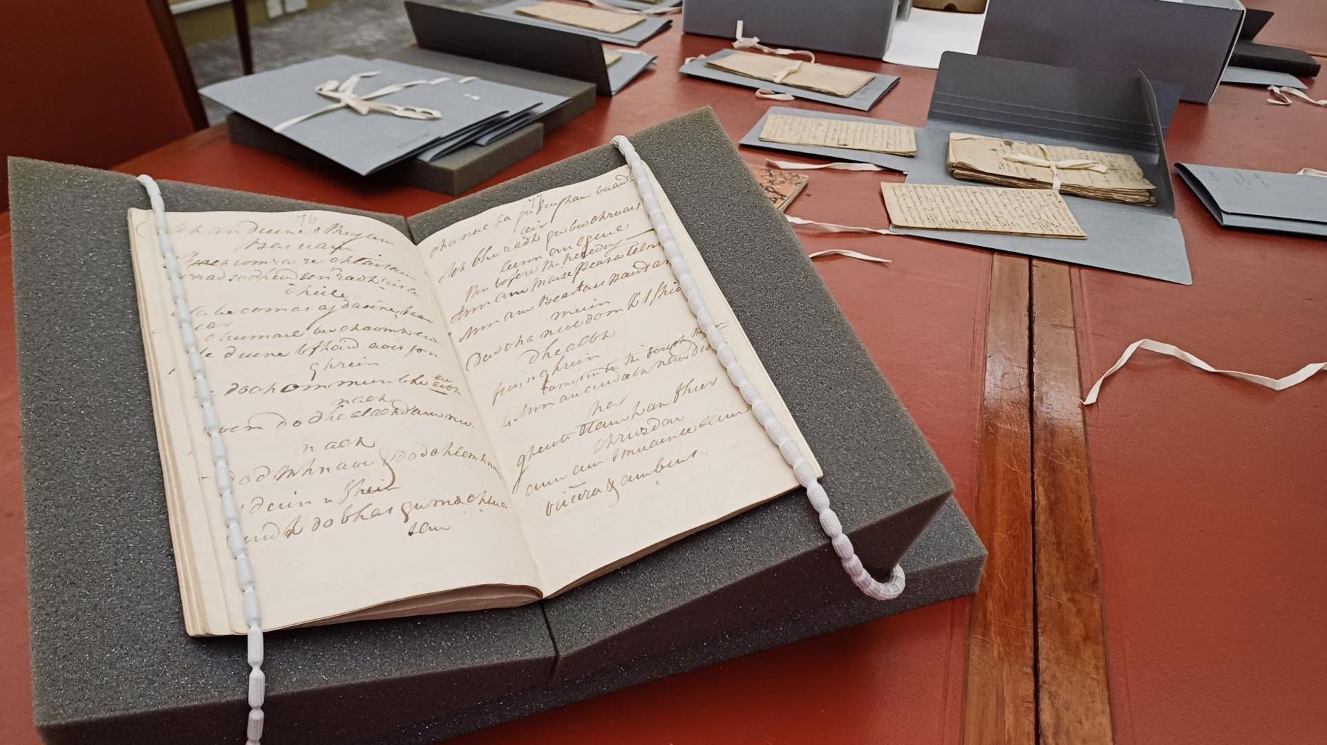 Pages of a manuscript open on a protective foam book holder. There are other manuscripts laid out on a red topped table.