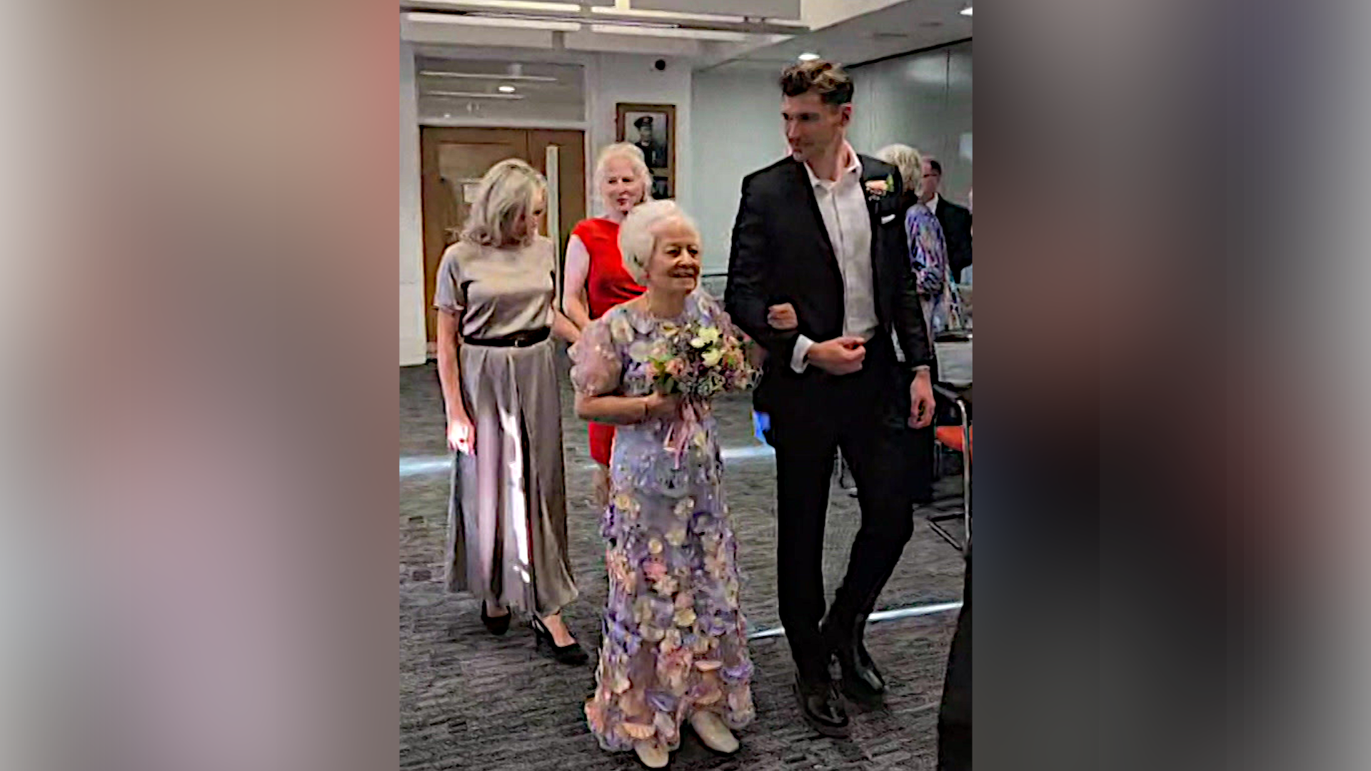 Jo is pictured walking down the aisle of a registry office with one arm looped in her grandson's. He wears a black suit with a white shirt underneath and he looks at his grandmother. Two women follow them behind.