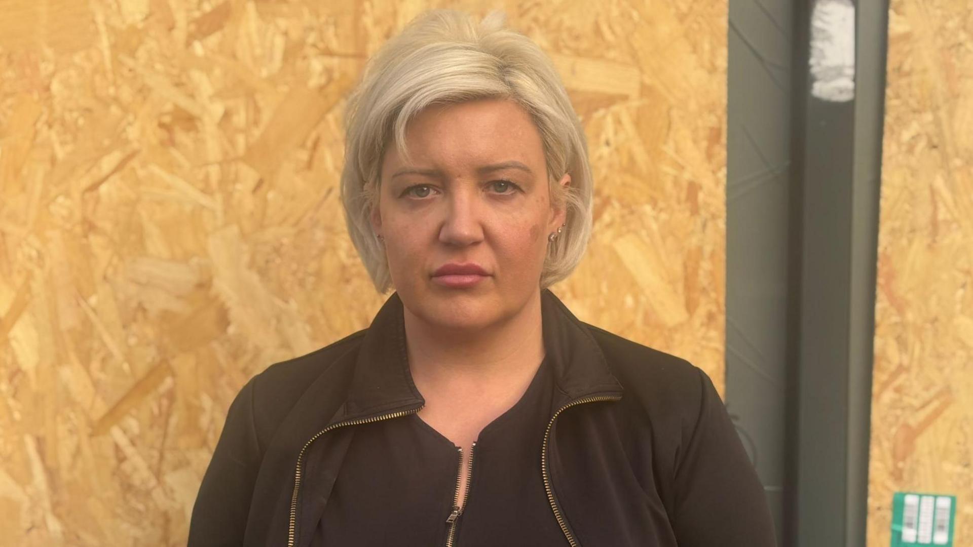 A profile image of Sarah Clay, with short blonde hair, wearing a black sweater in front of a boarded-up window. 