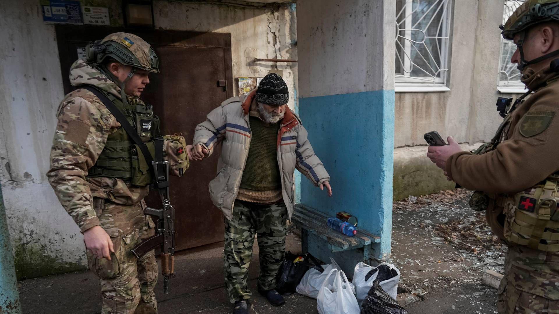 A bearded man leaves his home in Pokrovsk. Next to him on the ground are some possessions in carrier bags. An armed Ukrainian worker is holding his hand.