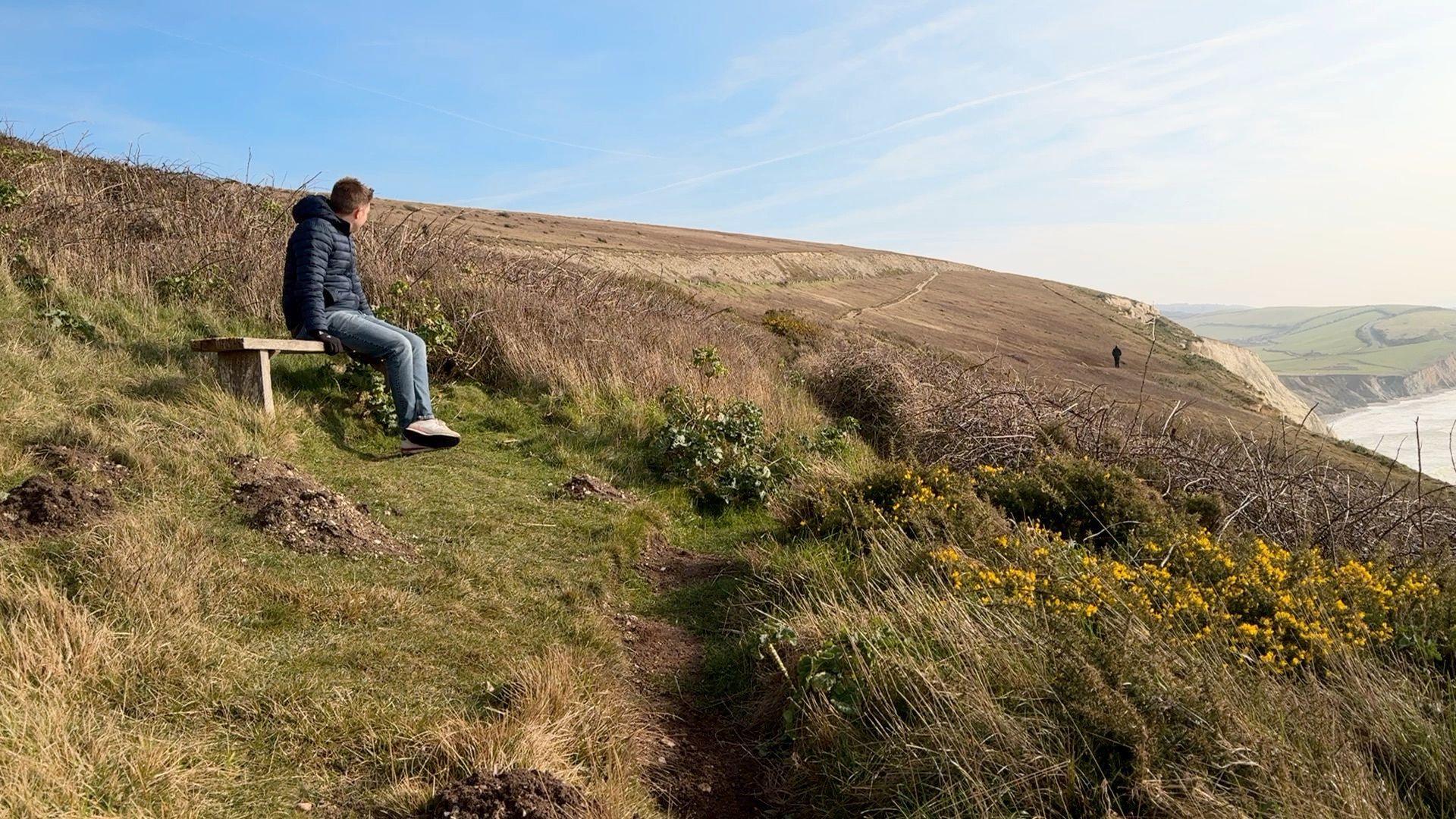 A man is sat on a bench on a hilltop. He is wearing a blue coat and blue jeans with white shoes. There is grass all around and the sky is blue. The land falls away where the cliffs cascade down to the sea below. 