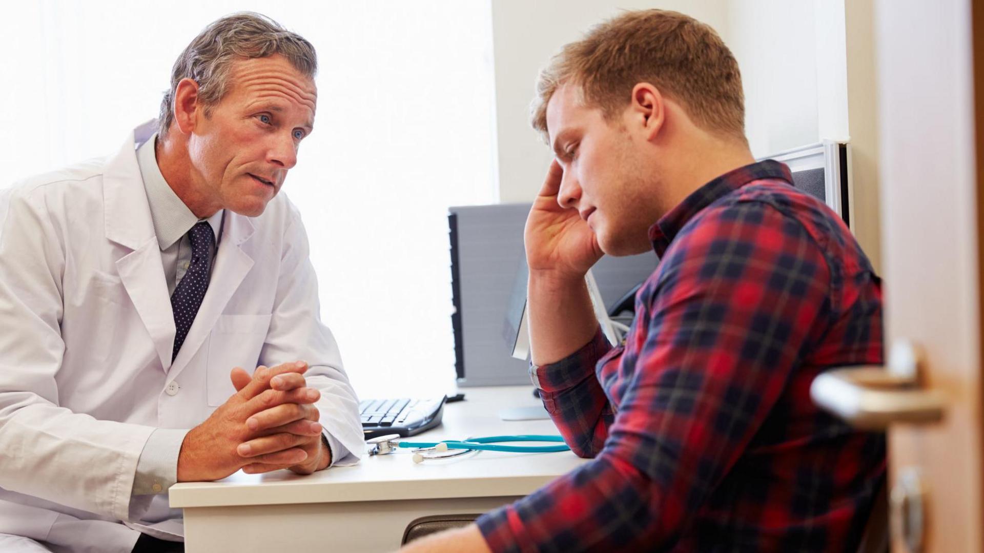 A doctor in a white coat talks to a sad looking young man in a checked shirt