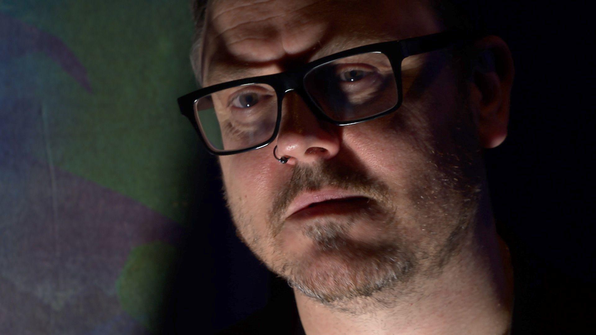 Ashley Thorpe of the Hell-Tor Festival, wearing dark rimmed glasses, with a nose ring through his right nostril, looking serious against a dark background