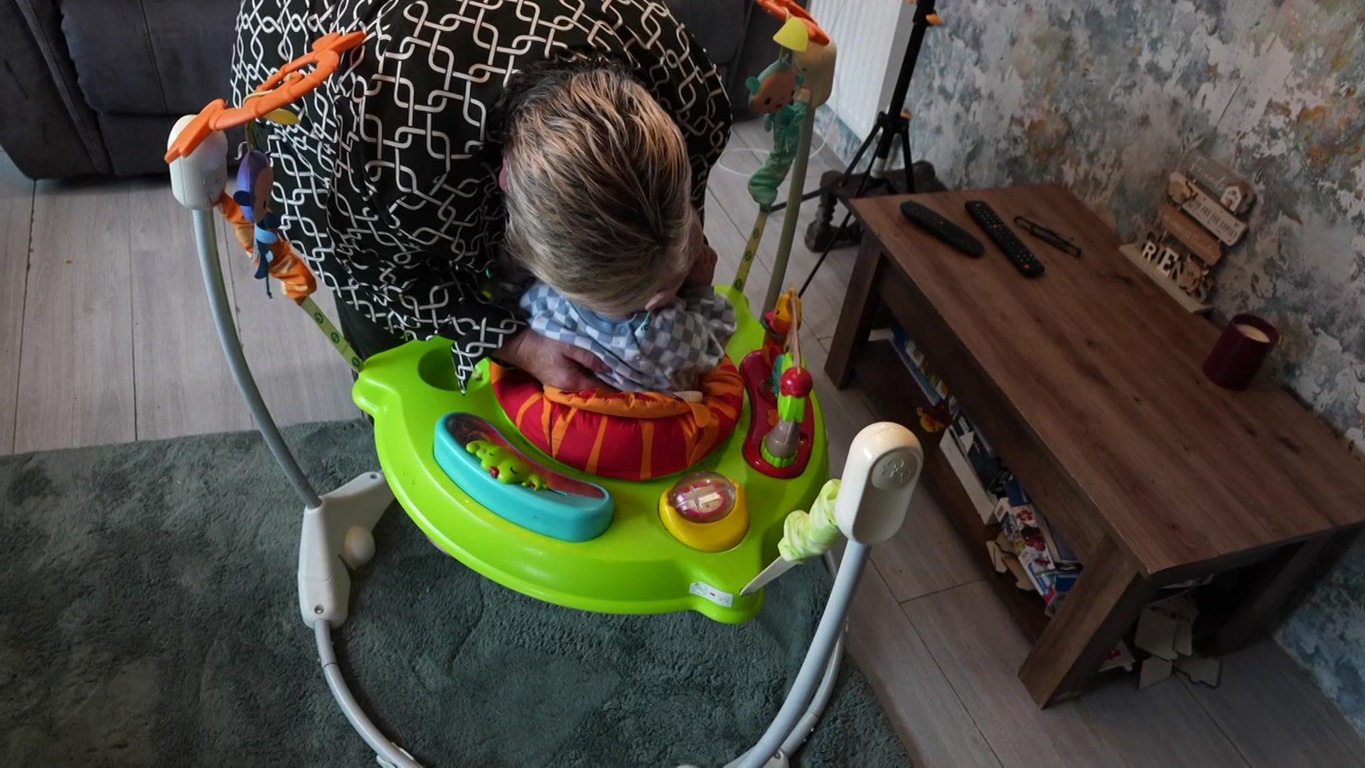 A baby is placed into a green bouncer in the middle of a living room in a static caravan
