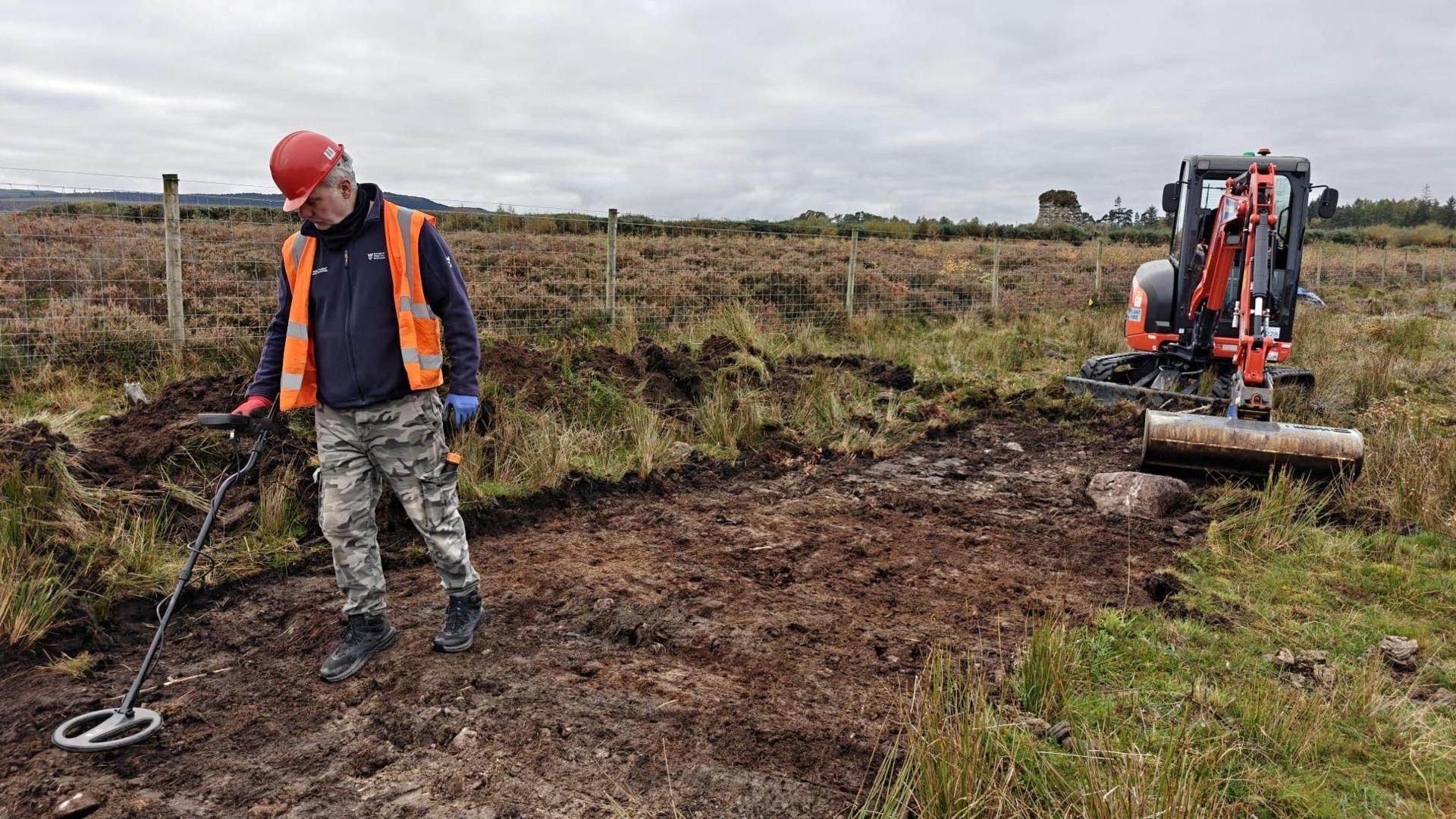 A man wearing a red hard hat, and a high visibility vest over a blue fleece, walks along an area of excavated ground with a metal detector. Behind him is a small red and black excavator. There is a low wire fence. On the other side of the fence is an area of heather-covered battlefield.