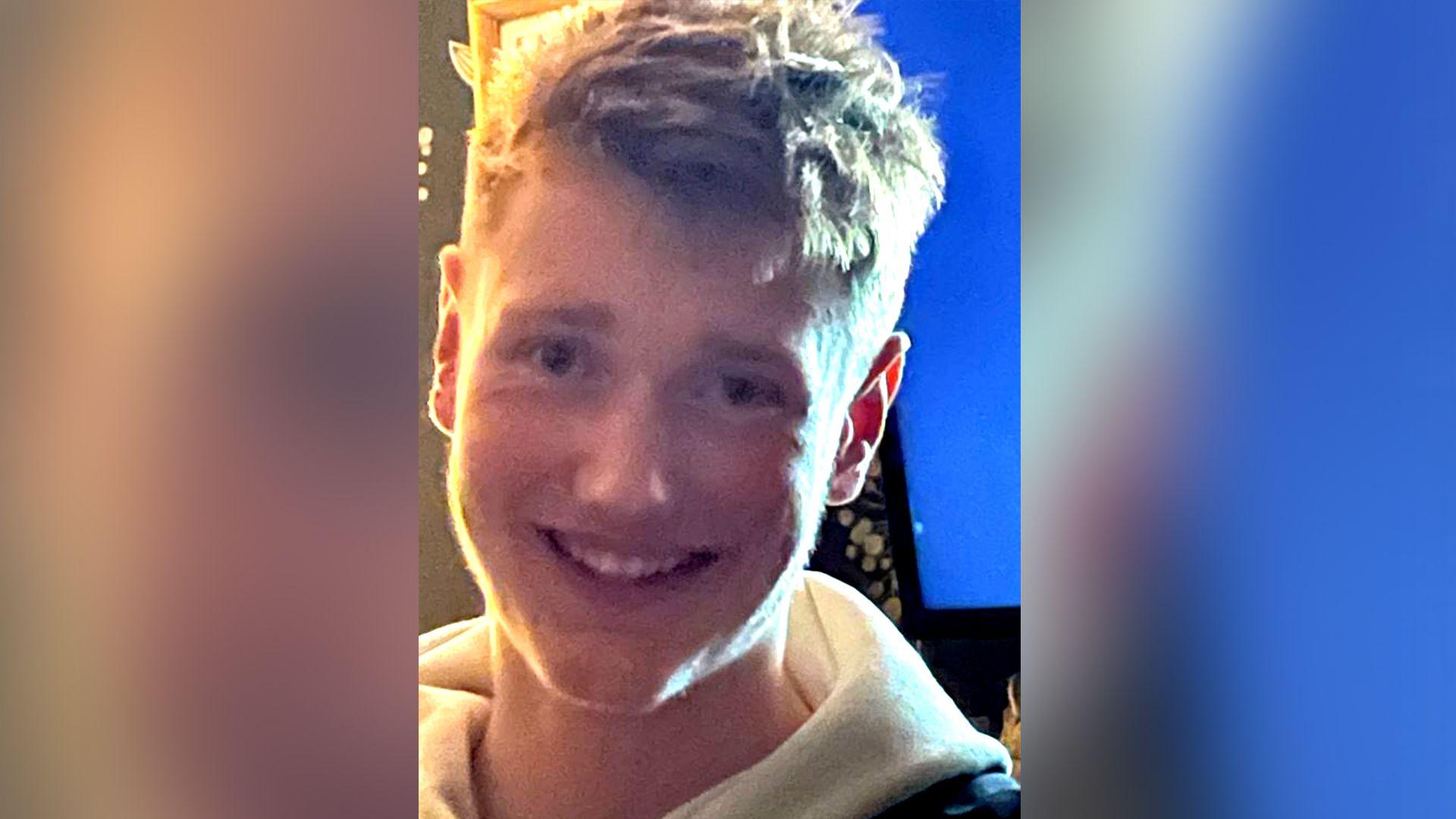 A young man with blonde hair smiles at the camera. He is wearing a hooded white top.