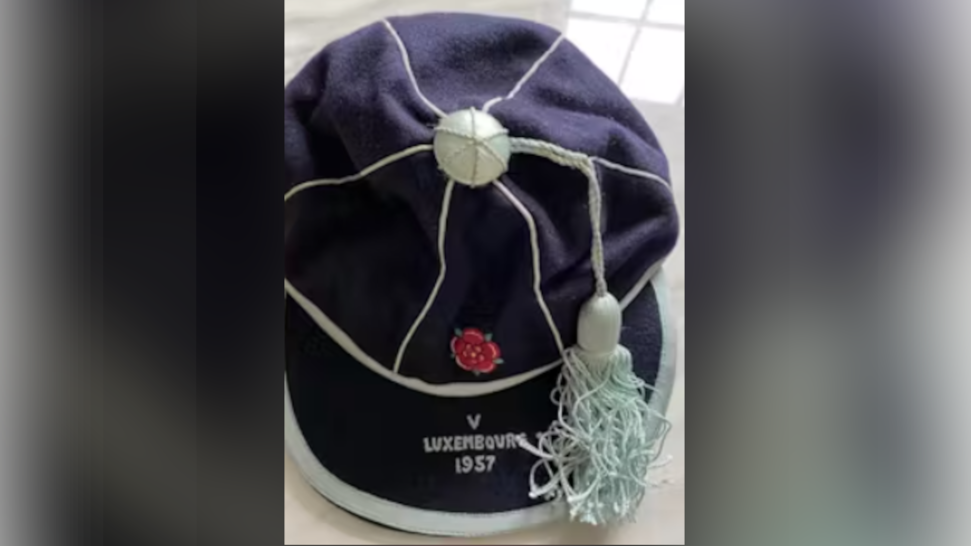 A blue cap with white details on it and a light blue tassle rests on a white surface. The English rose is embroidered on the front along with the words: V Luxembourg 1957