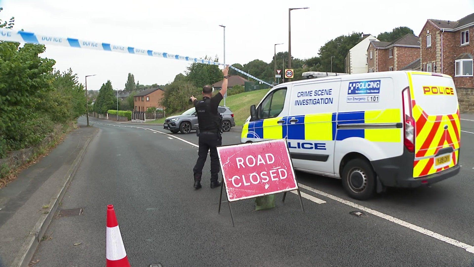 A CSI van going through a road closure. A police officer is lifting police tape to allow the van through. There is a road closed sign in the left-hand lane.