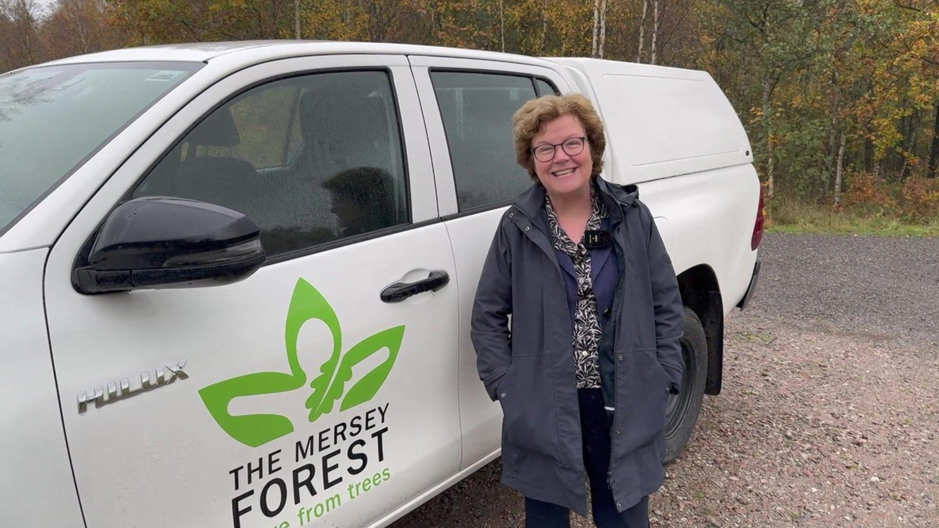 Professor Eunice Simmons has short brown hair and is wearing dark rimmed glasses and a blue coat and is stood next to a white van with The Mersey Forest logo on.