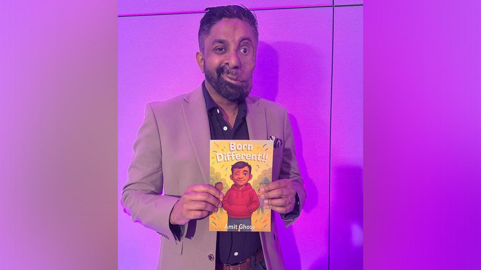 A man wearing a beige jacket and blue trousers stands in front of a purple backdrop holding a book entitled Born Different.