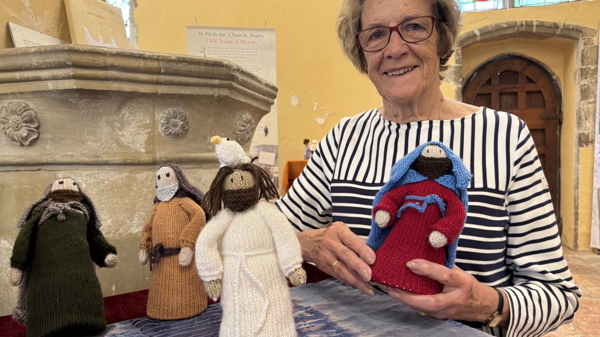 Jane Freeman, wearing a black and white striped top, stands by a church font and holds a knitted biblical figure of a man. Three other figures are stood on a table in front of them, one with a white dove depicted on its head.