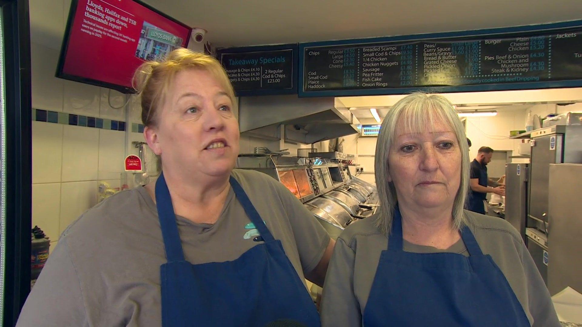 Two women standing in a fish and chip shop. There is a man in the background in the kitchen. 