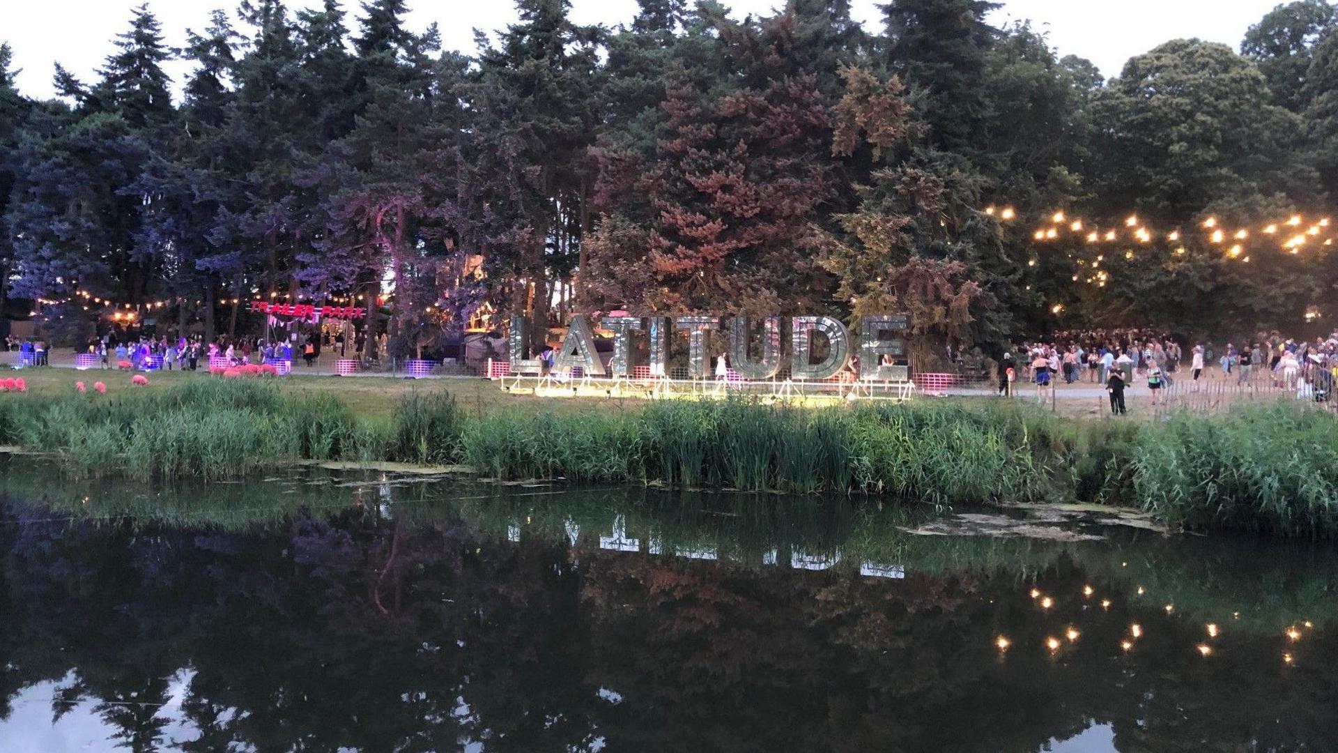 A river bank with a large mirrored sign saying "Latitude". There are pink sheep and lights in the trees. The river is in the foreground.