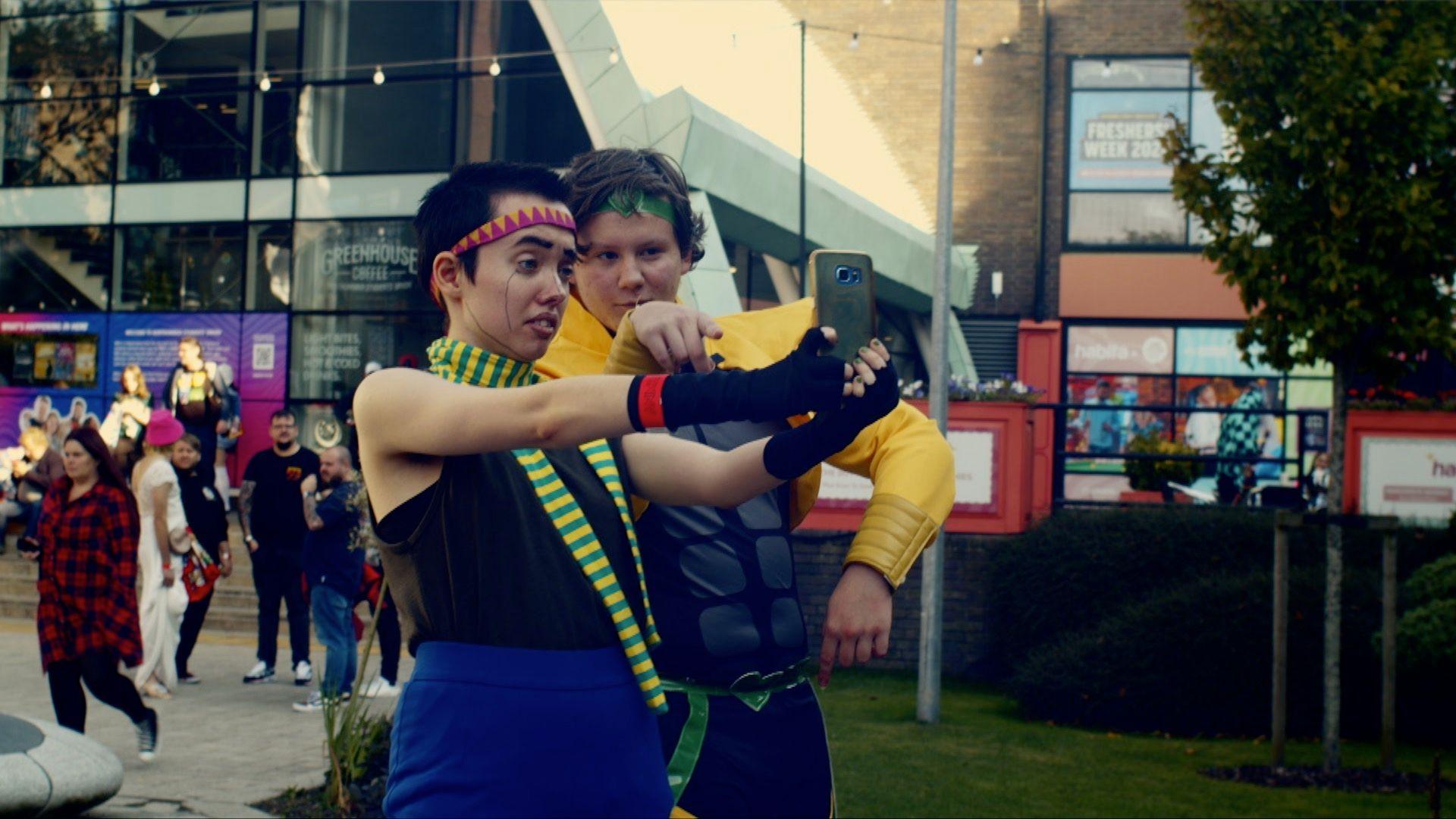 Chris is dressed in a yellow outfit and is having a selfie taken by a woman in Anime cosplay. They are stood outside, with a small crowd behind them.