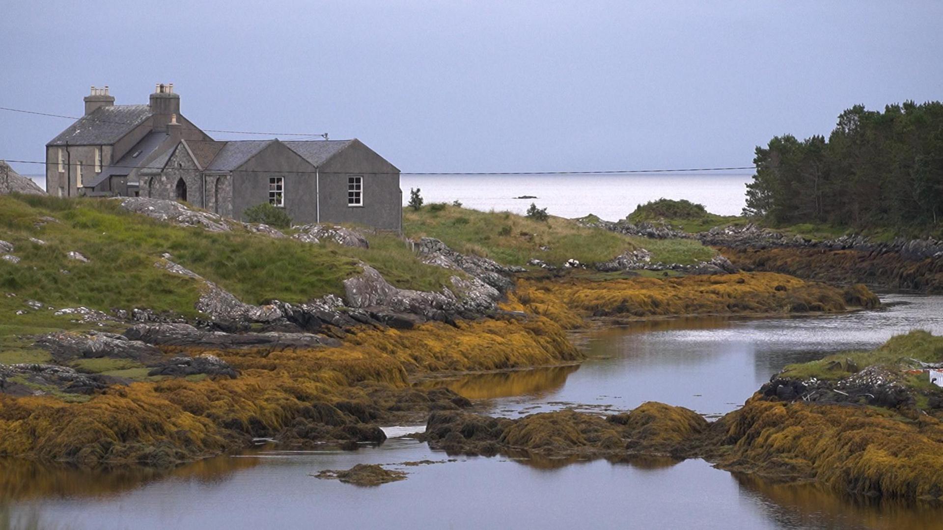 Sealladh de dh'eaglais ghlas agus togalach glas ri a taobh.  Tha iad gu math faisg air a' chladach agus chìthear muir agus feamainn odhar agus creagan.  Tha coille bheag ann cuideachd. 