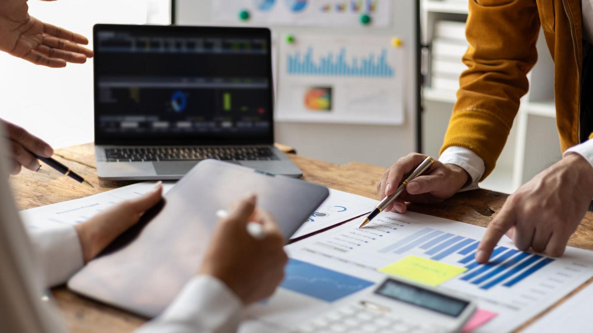 Work colleagues reviewing charts and graphs on a desk in an office. One is standing up pointing at a chart, while another person is sitting down reading a tablet.