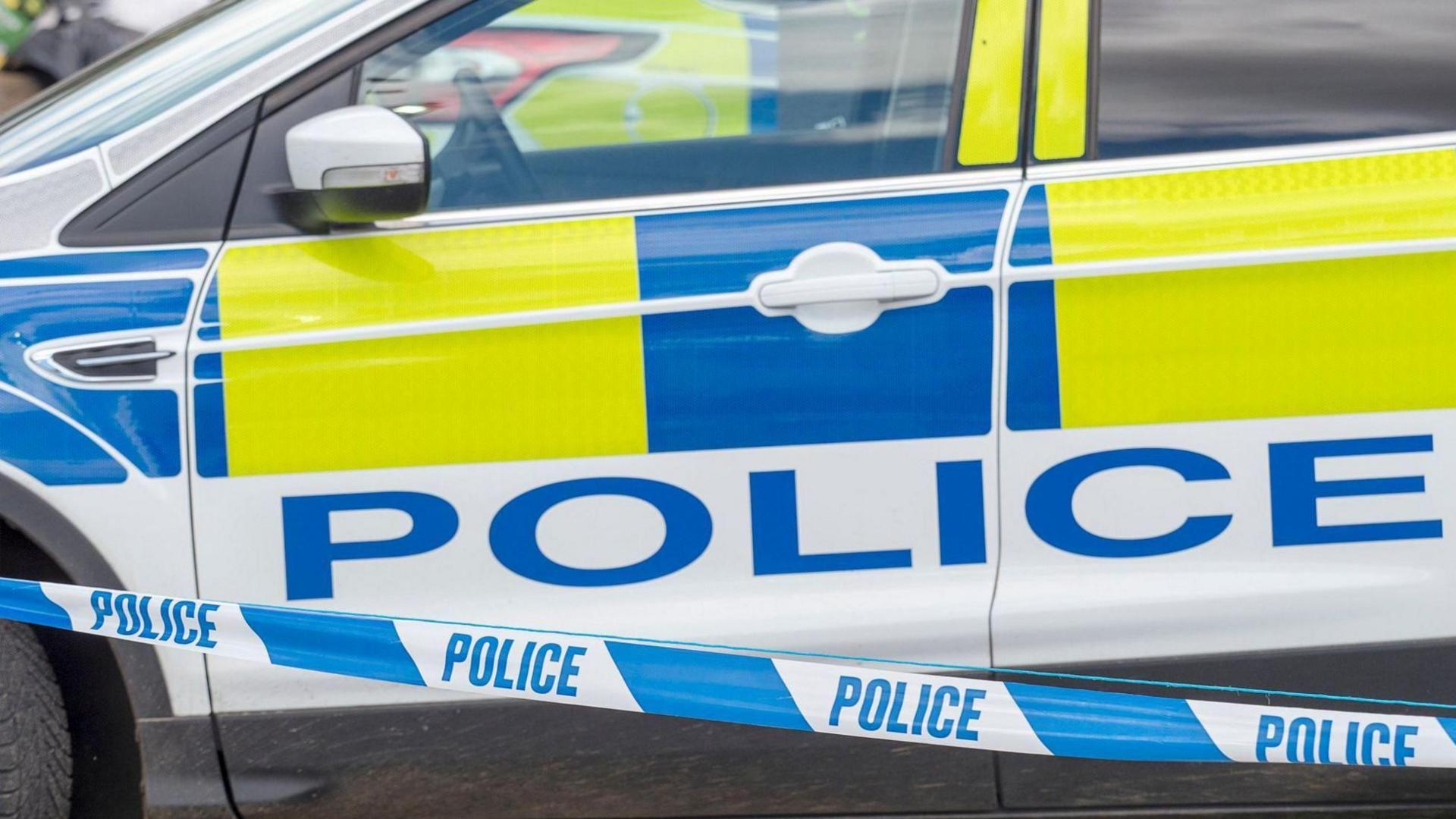 A stock image showing a parked police car with hi-vis blue and yellow sticker decals. In the foreground there is blue and white police cordon tape.