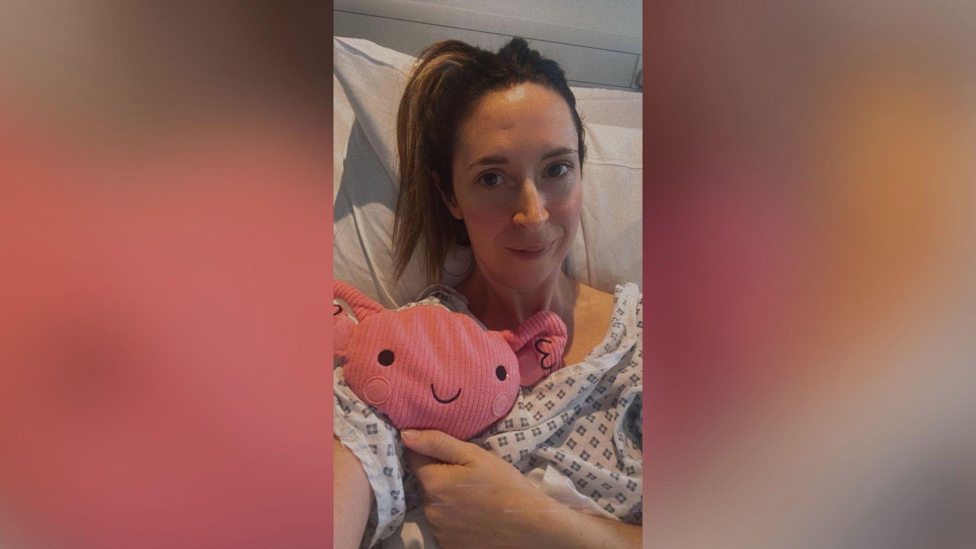 Gynaecology patient Claire lies in her hospital bed after surgery with a womb-shaped toy as her mascot.