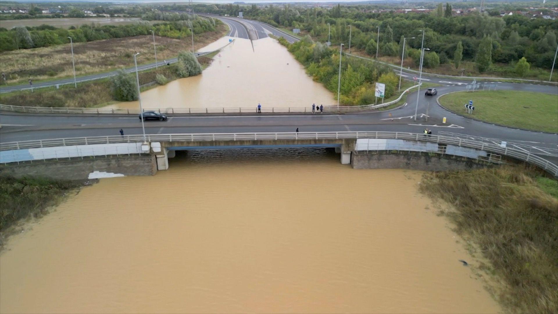 Brown water has submerged the A421 as it runs under a bridge. The road is almost impossible to see until the water stops in the horizon and the road continues.