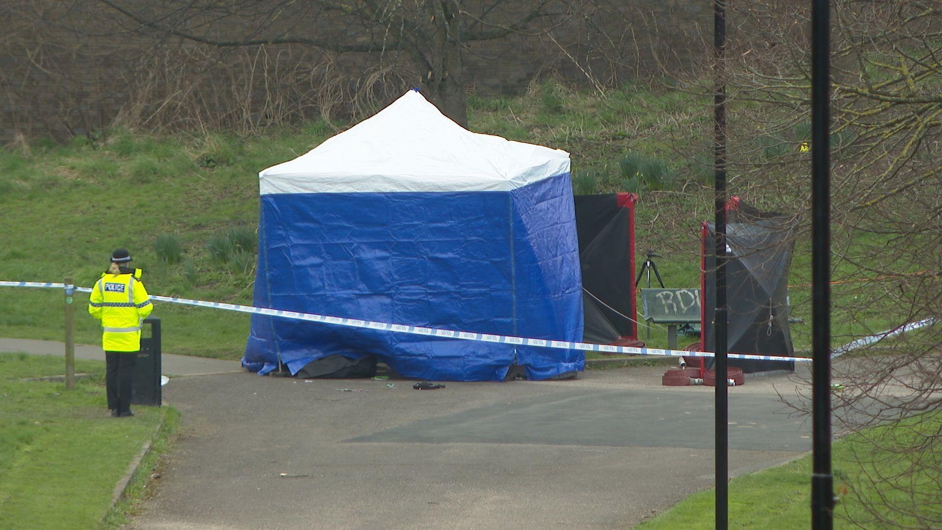 A blue police tent on a pathway surrounding by blue and white tape. A police officer in a yellow high-viz jacket stands nearby. There are two black privacy shields next to the tent.