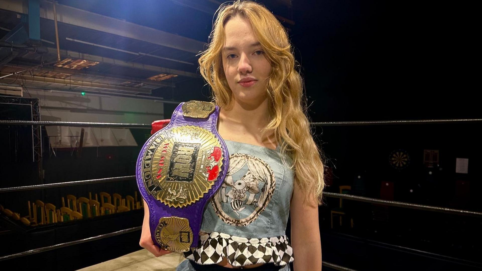 Annabelle with long blonde hair holding her purple and gold European Bellatix belt over her right shoulder. 