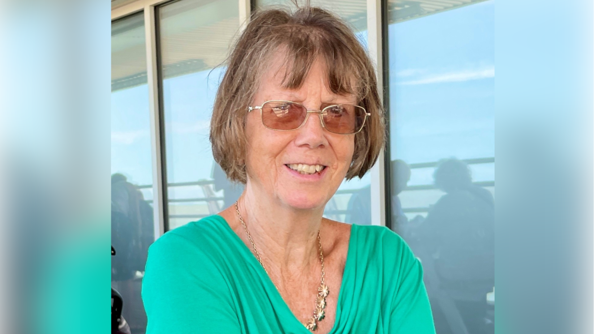 Christine Murton is in front of a large glass window. She has short brown hair and is wearing a green outfit with a gold statement necklace and glasses. She is looking directly at the camera and smiling.