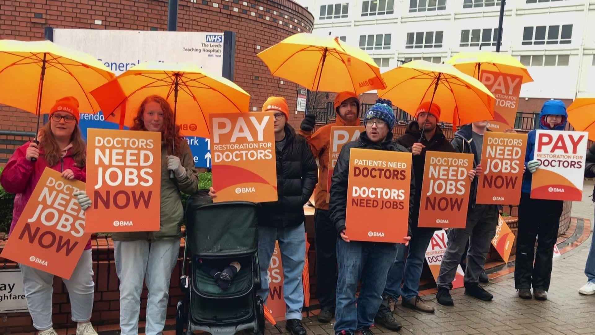 A group of people holding orange placards and umbrellas. The placards read 'Doctors need jobs now'.