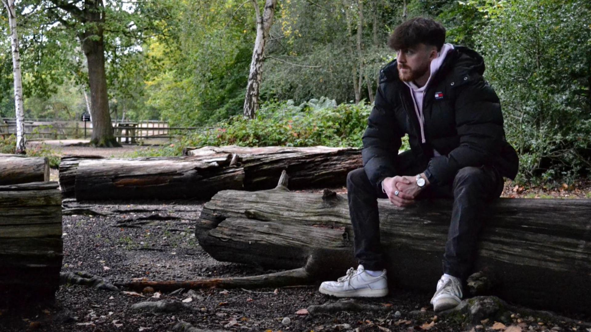 Connor Scott with short brown hair and short brown beard, wearing blue jacket and trousers with white trainers, sitting on a tree trunk with trees in the background