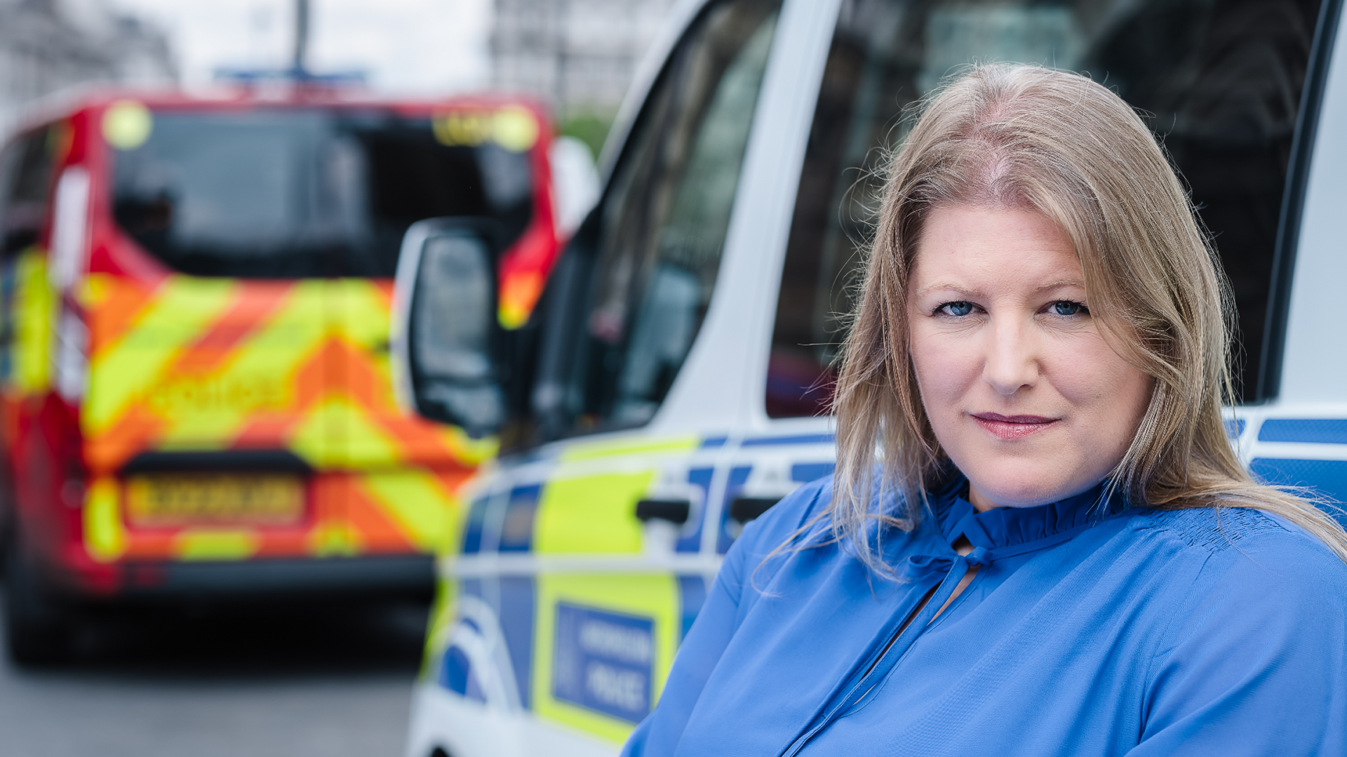 Donna Jones is pictured standing in front of two police vehicles. She has long, straight, light brown hair and wears a blue blouse.