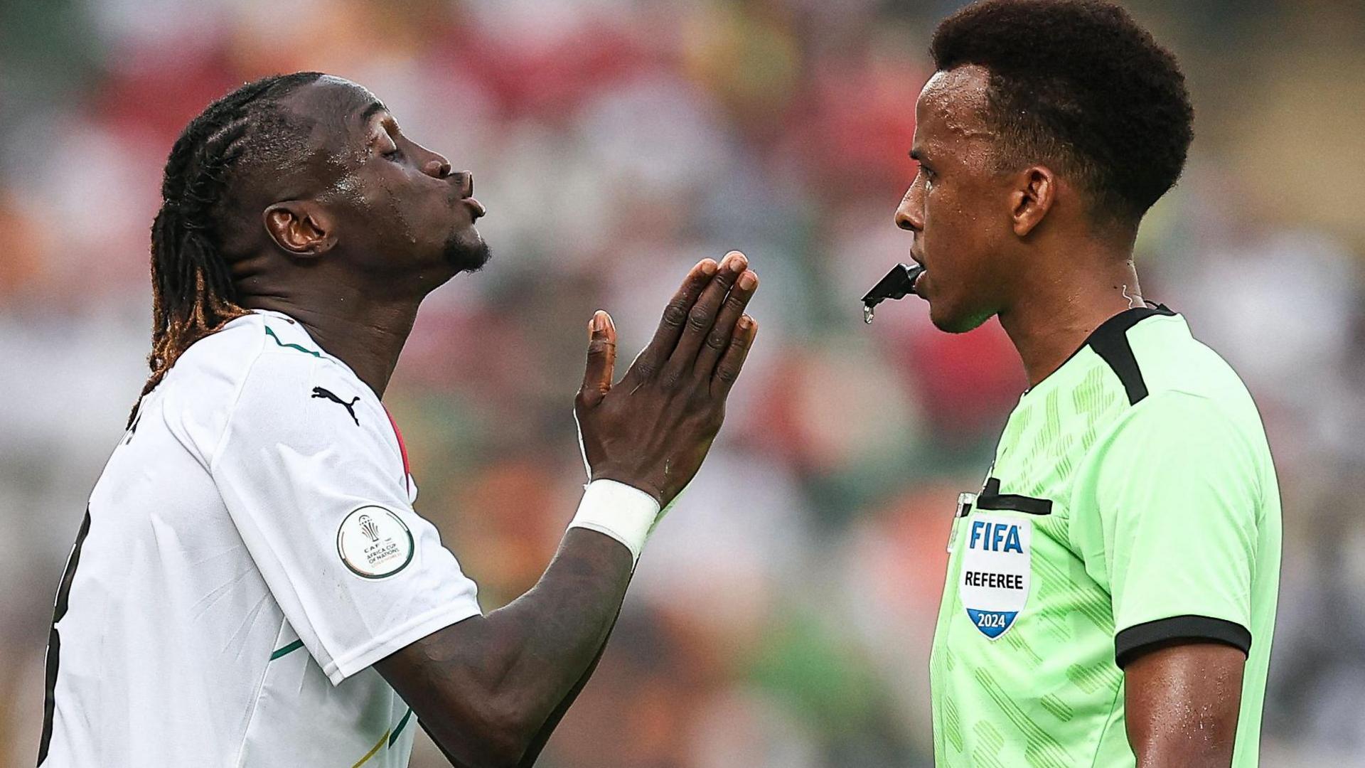 A player wearing a white football shirt holds his right hand up in front of him as he pleads with referee Omar Artan, who holds a whistle in his mouth as he wears a bright green referee's shirt