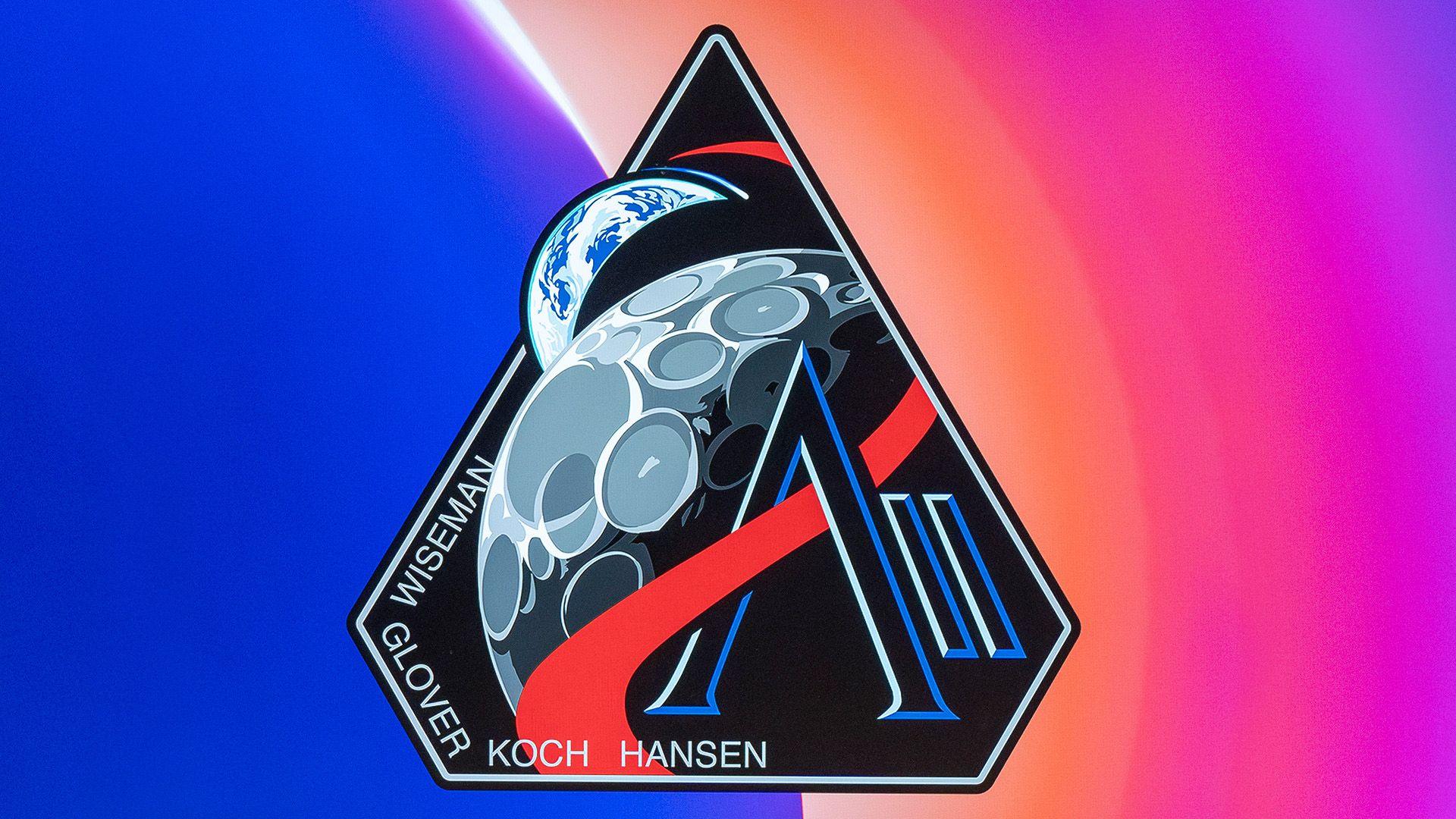 The emblem is the shape of an upside down diamond. Its background colour is black and has a grey line around the edge. In the bottom left hand corner of the patch are the names Wiseman, Glover, Koch and Hansen. In the middle of the patch is a cartoon moon. There is a red wave line going over it. It also goes through a black and dark blue AII. Behind the moon and sticking out of the diamond shape is a cartoon image of the Earth