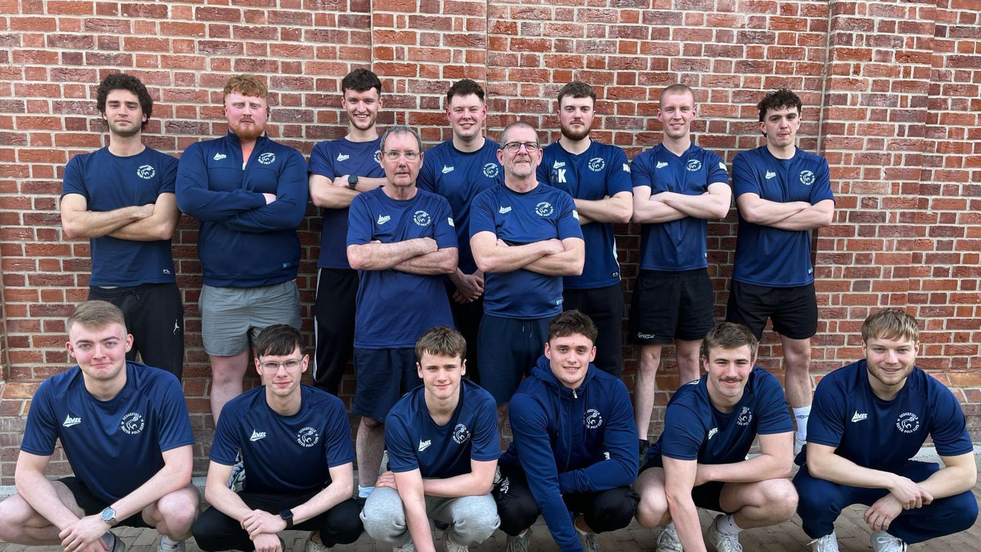The full water polo team posed in front of a brick wall with a row crouched down in front of a row standing up.