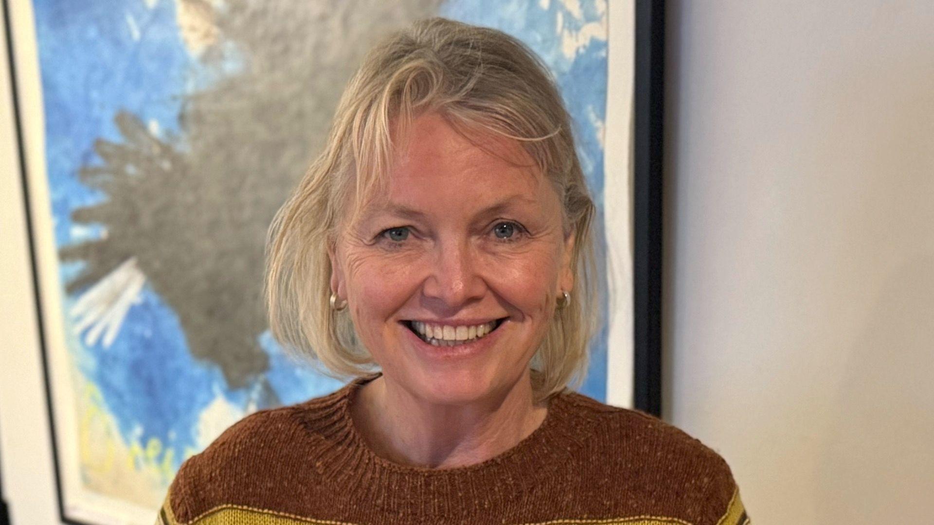 A woman - Sherri Singleton - smiling straight at the camera. She is wearing a brown knitted jumper and is standing in front of a wall mounted painting.