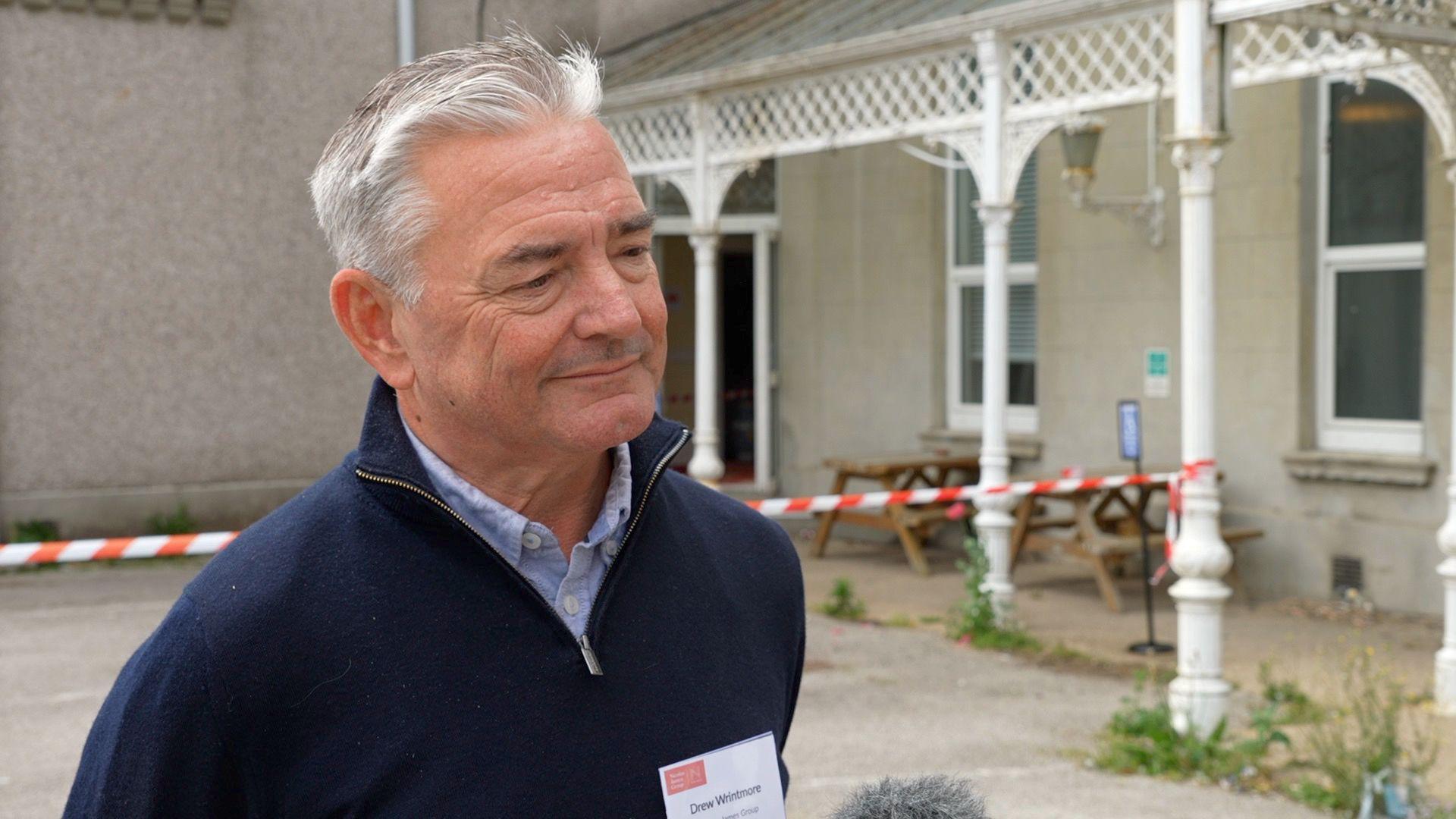 Drew Wrintmore stands outside a building, wearing a dark blue sweater and a name tag. The background features white decorative columns, windows, red and white caution tape, and picnic tables.