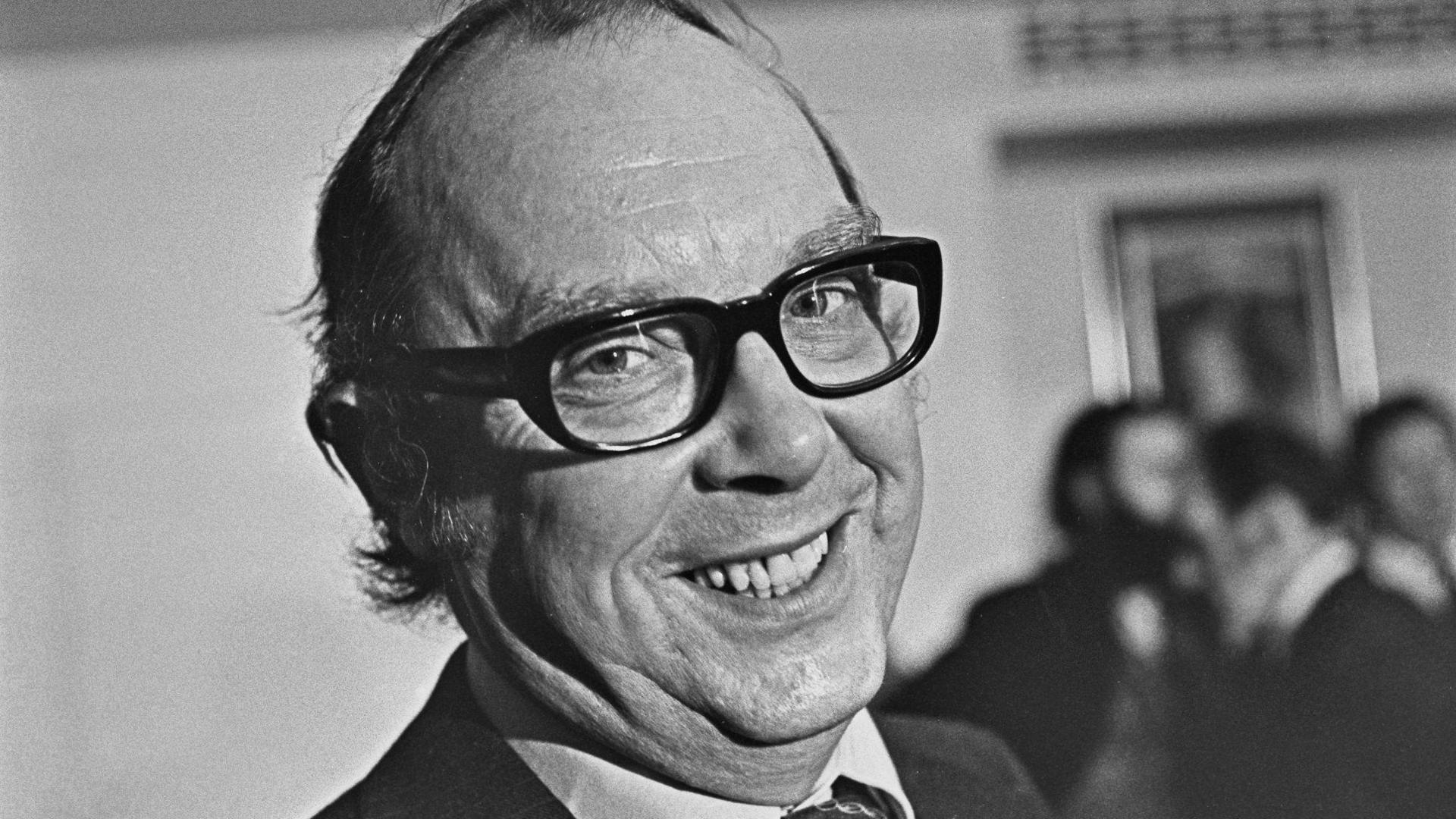Close up of Eric Morecambe (1926 - 1984) of comedy double act Morecambe and Wise, who is wearing black glasses and smiling. The photograph is in black and white and there is a blurred image of people stood behind Eric.
