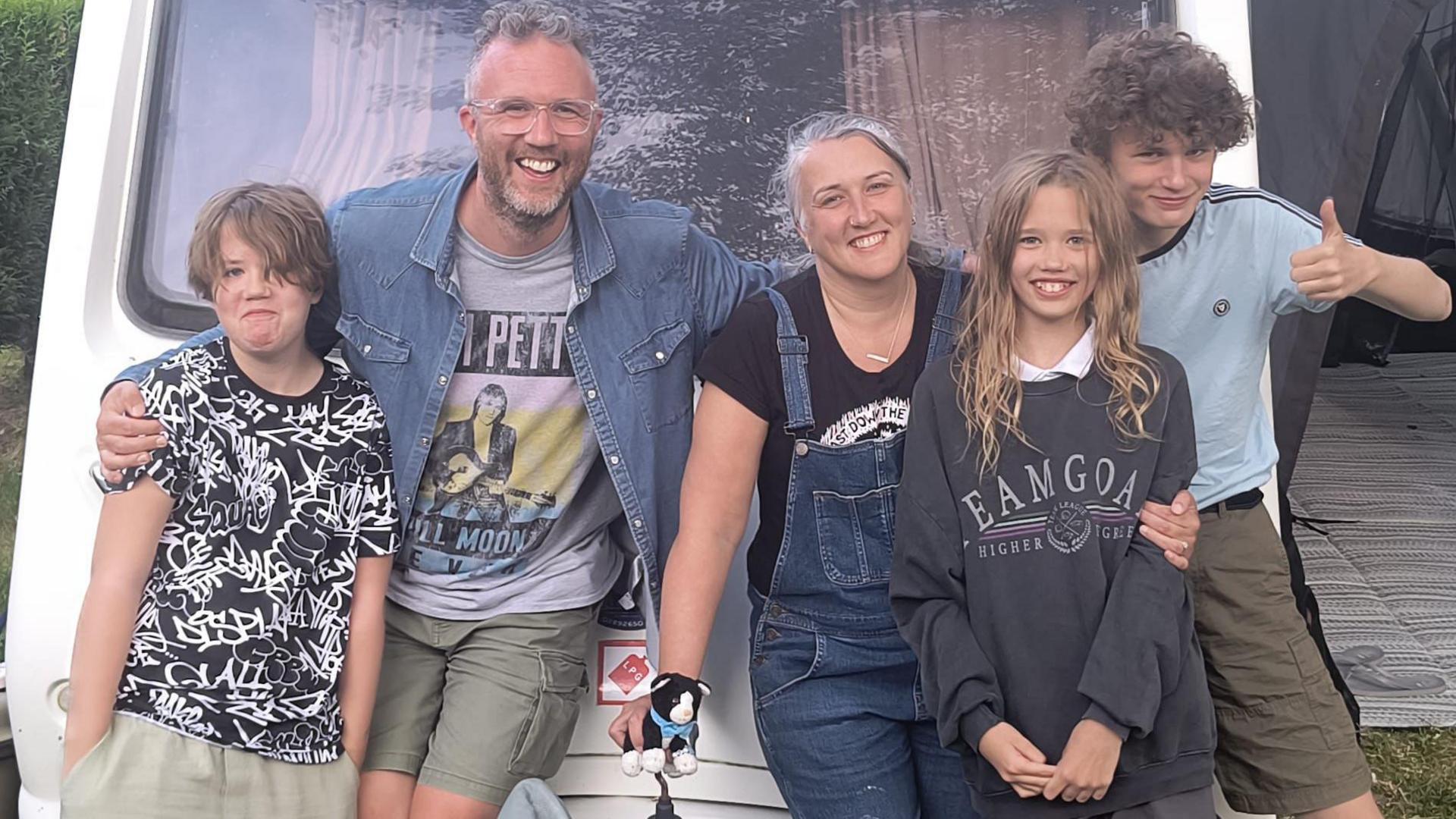 Five people standing in front of a caravan with arms round each other. On the left a child is wearing a black t-shirt with white graphic writing across it, they have brown hair. Next a man wearing glasses with short grey hair. he is wearing a denim shirt with a graphic t-shirt underneath and green shorts. Next a woman is wearing denim dungarees with a black t-shirt. she has grey hair tied up. next a child is wearing a large sweatshirt and has long blonde hair. Last a child is wearing a bright blue t-shirt and has curly hair, he is giving a thumbs up. 