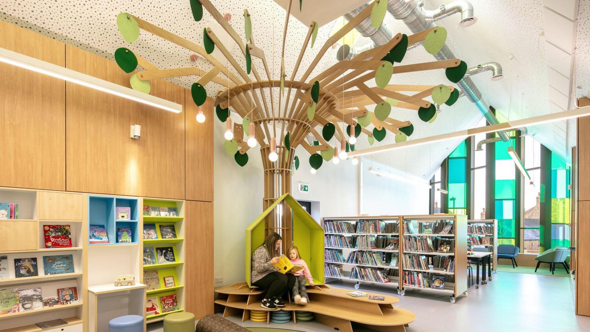 A woman and a child sit on a bench shaped into a tree. Books can be seen on the left wall and arranged on shelves at the back of the room.