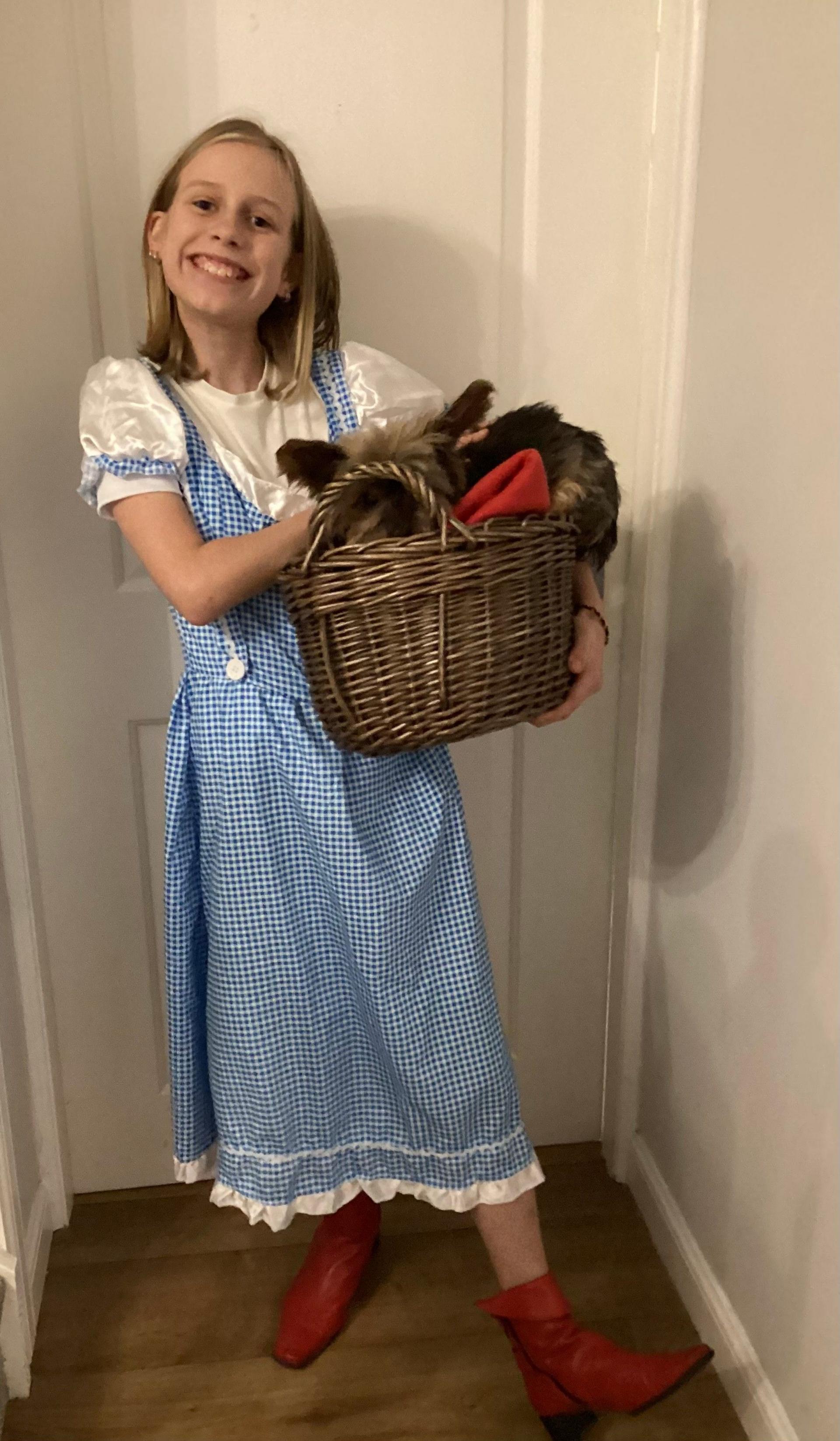 Hannah stands with a blue check dress, red boots and a basket with a dog in