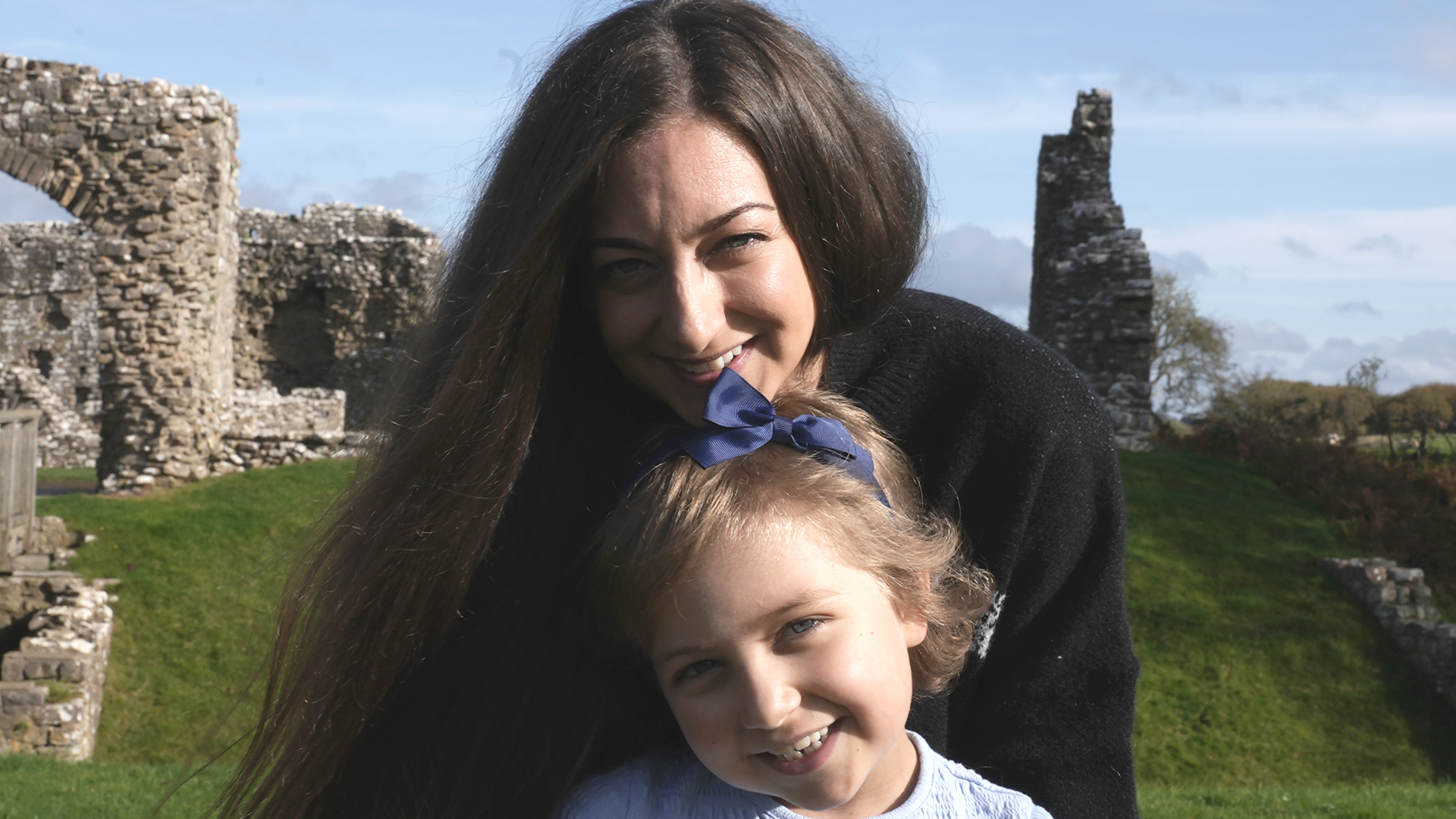 Charlotte and Betsy in the ruin of Ogmore Castle. Charlotte is embracing Betsy from behind. Charlotte has long dark hair and is wearing a black jumper. Betsy has a navy ribbon in her short fair hair and is wearing a pale top. 