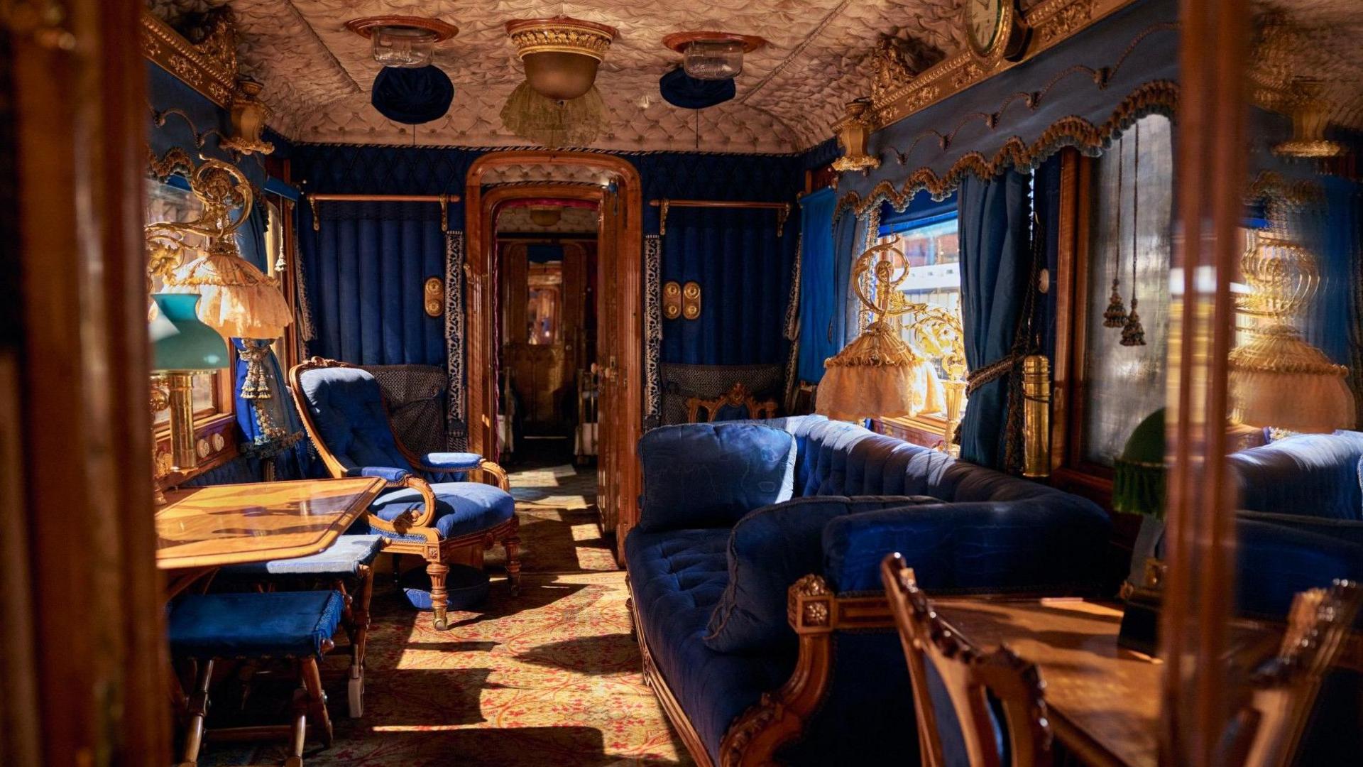 The interior of a royal rail carriage designed and used by Queen Victoria. The furnishings are made of wood and plushly upholstered in a dark blue velvet and include a writing desk, couch and armchair