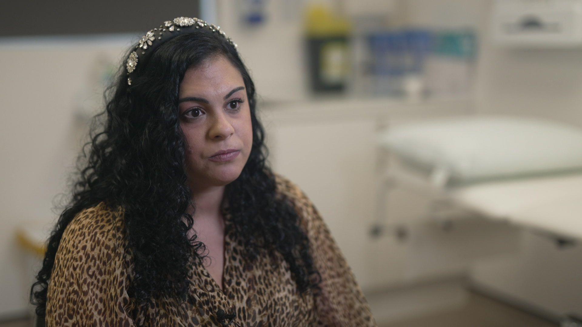A woman wearing a leopard-print shirt sat in a doctor's room. A white doctor's bed is in the background. She is sat down, looking past the camera and out into the distance.