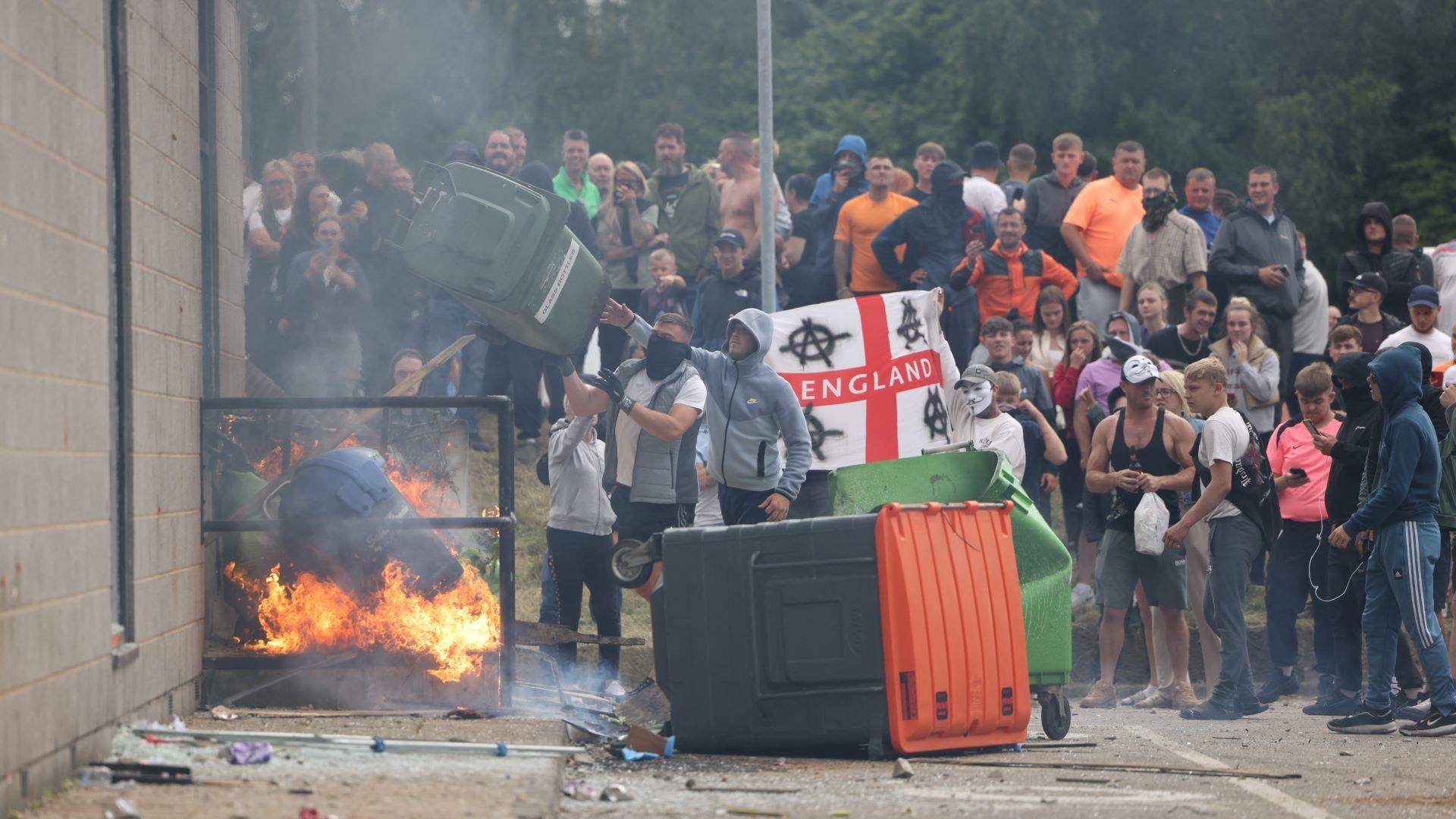 A bin is on fire while another bin is turned on its side. A man wearing a grey hooded top is throwing a green wheelie bin into the fire. An England flag with far-right slogans is held by some of the protesters.