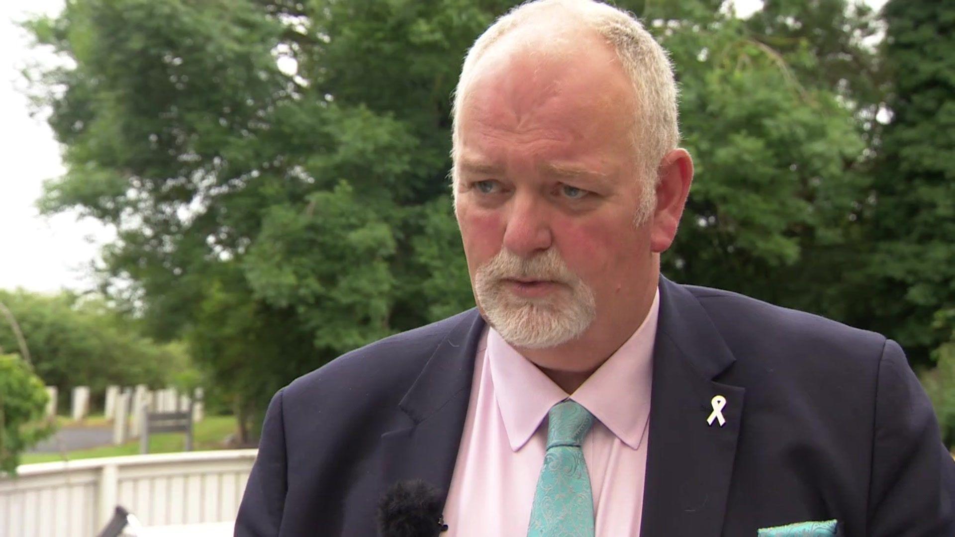 Jonathan Evison with a goatee-style beard standing in front of trees wearing a dark jacket, a pink shirt and a green tie