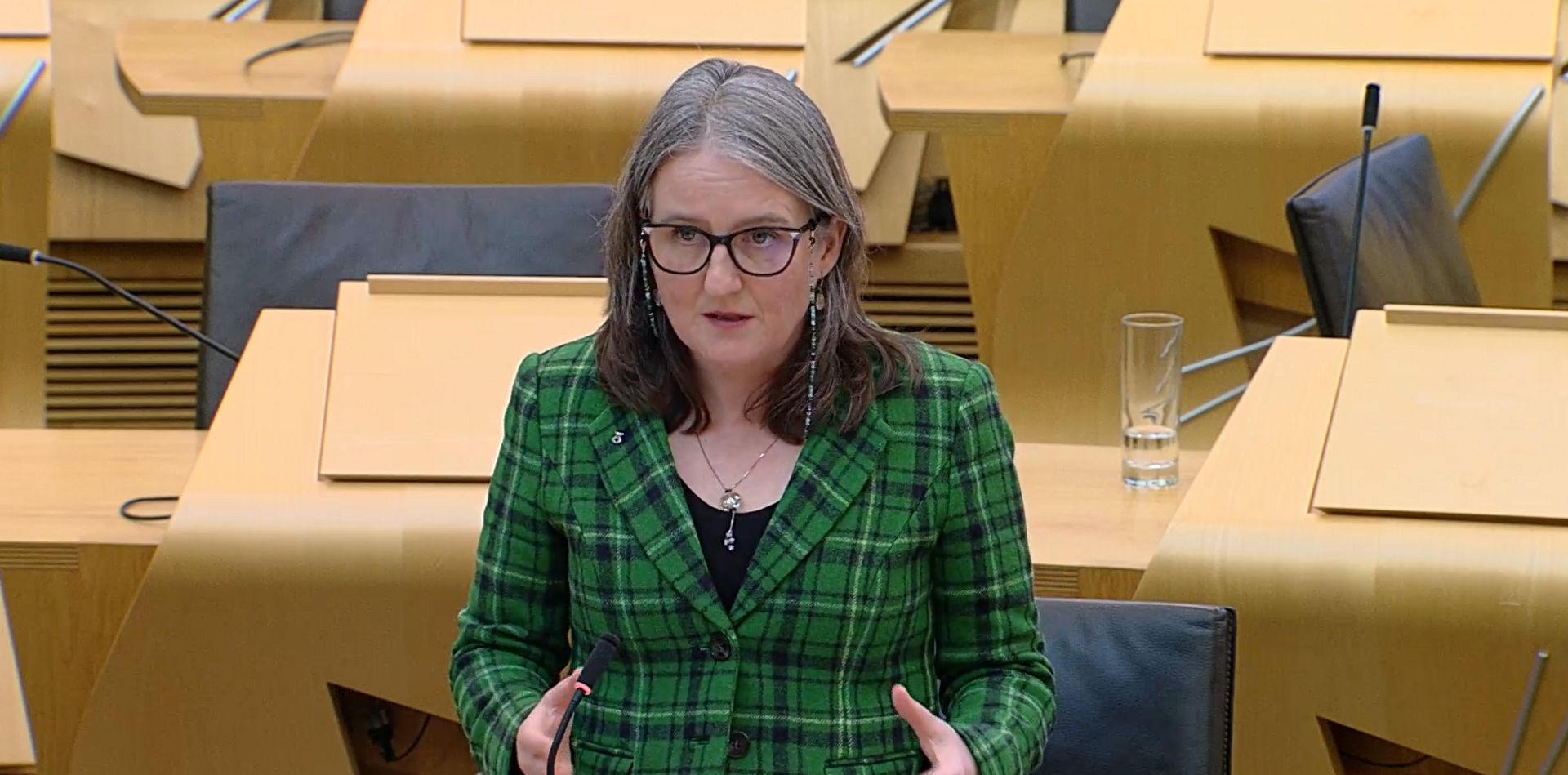 Maree Todd wears a green tartan jacket, black top, silver necklace and black-framed glasses with a retainer chain. She is standing in the chamber in Holyrood, with wooden desks and black seats behind her.