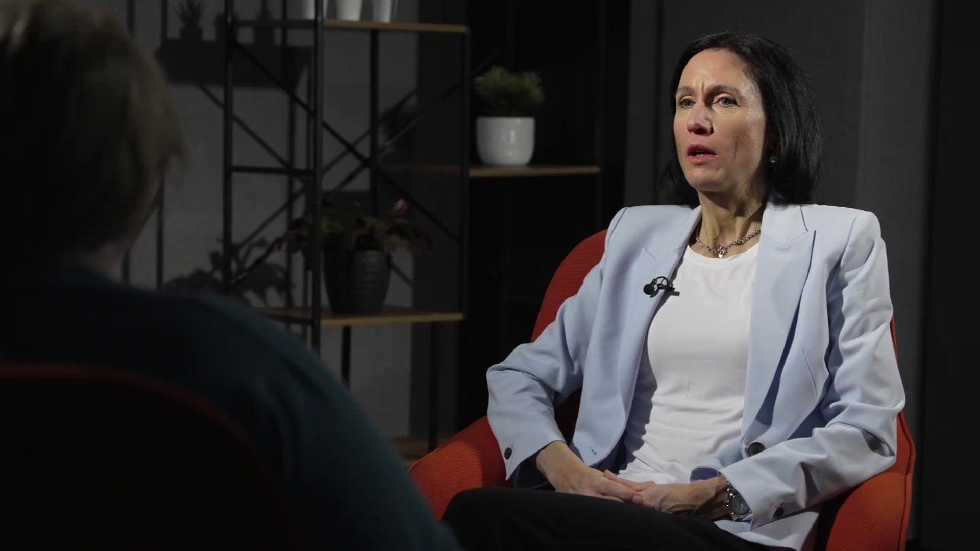 BBC reporter Fiona Trott, wearing a white shirt and blue jacket with long dark hair, sits in a chair facing a woman who is obscured to us. She has her back to the camera and is wearing a green top and has long brown hair. In the background are some wooden and metal shelves with pot plants on them.