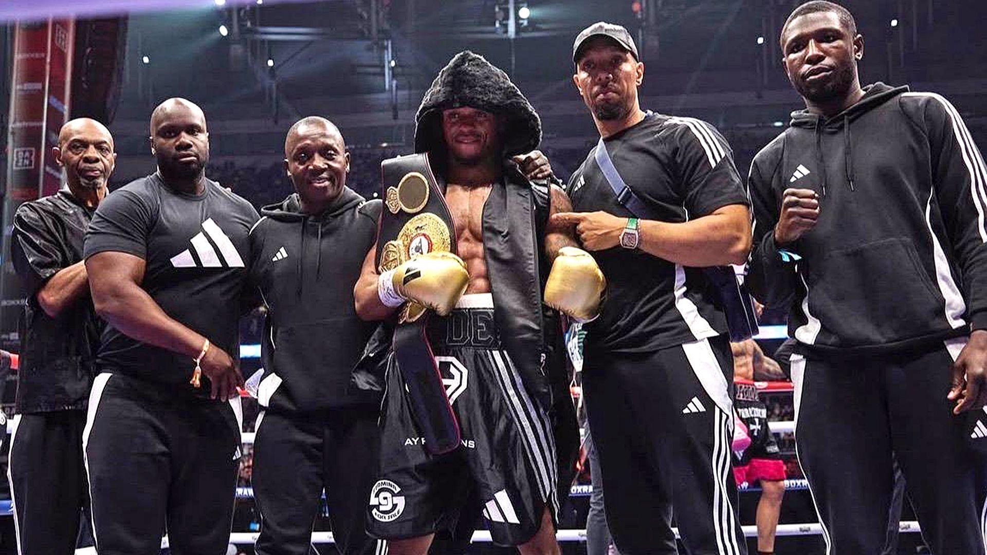 James Cook alongside Anthony Yarde and his team in a boxing ring after a fight. The team are wearing black tracksuits. Anthony Yarde, in the middle, has a black hood up and gold boxing gloves. He also has a title belt over his shoulder. 