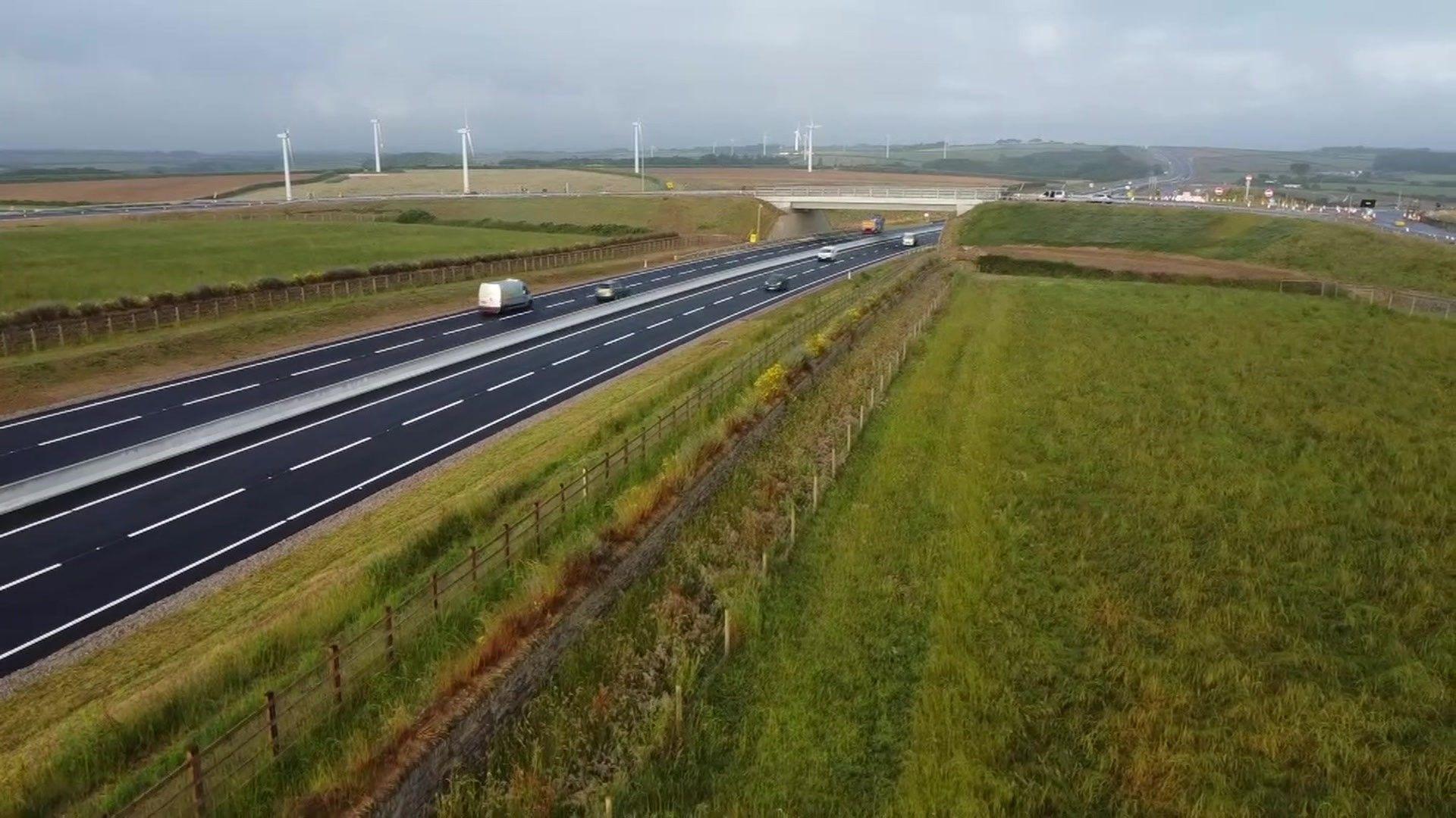 An aerial photo showing a stretch of the A30. There are wind turbines in the distance and a bridge across the dual carriageway. There are fields both sides of the road.