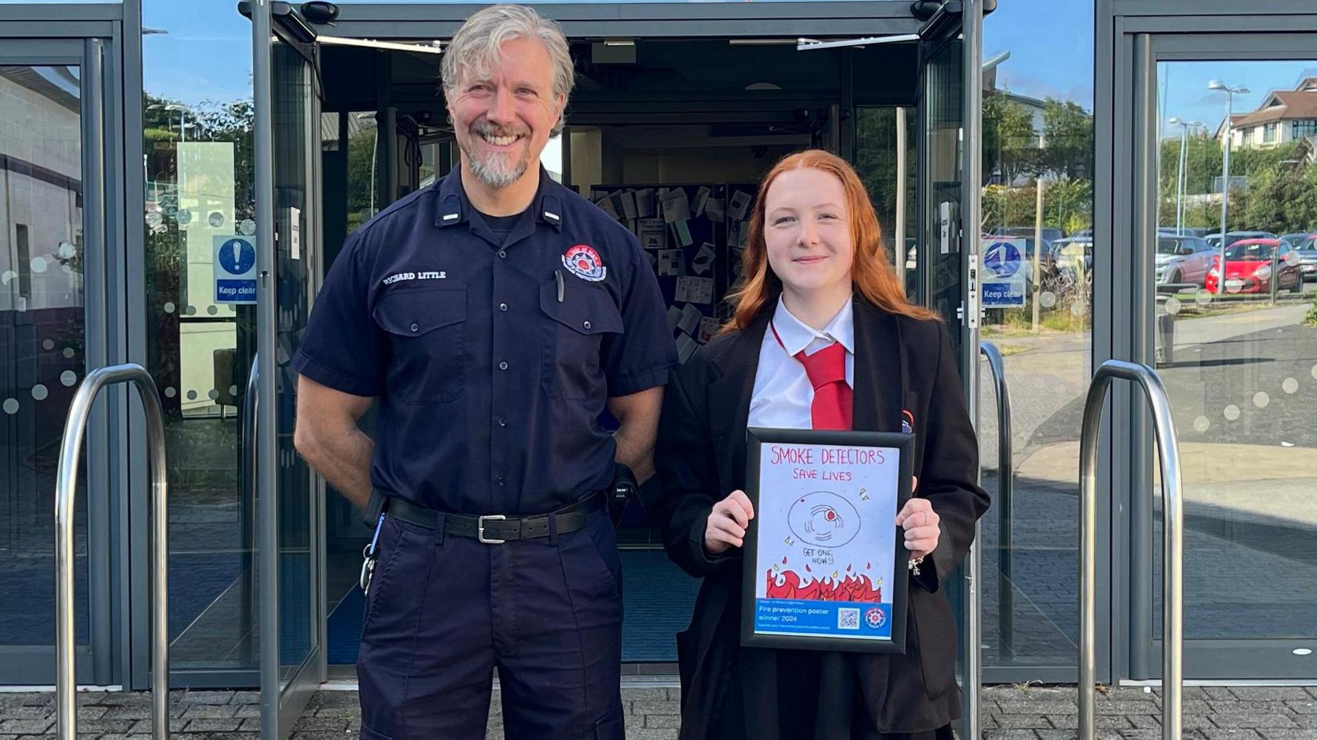 A blonde firefighter stood next to a ginger secondary school girl smiling holding her poster in front of St Ninian's High School entrance.